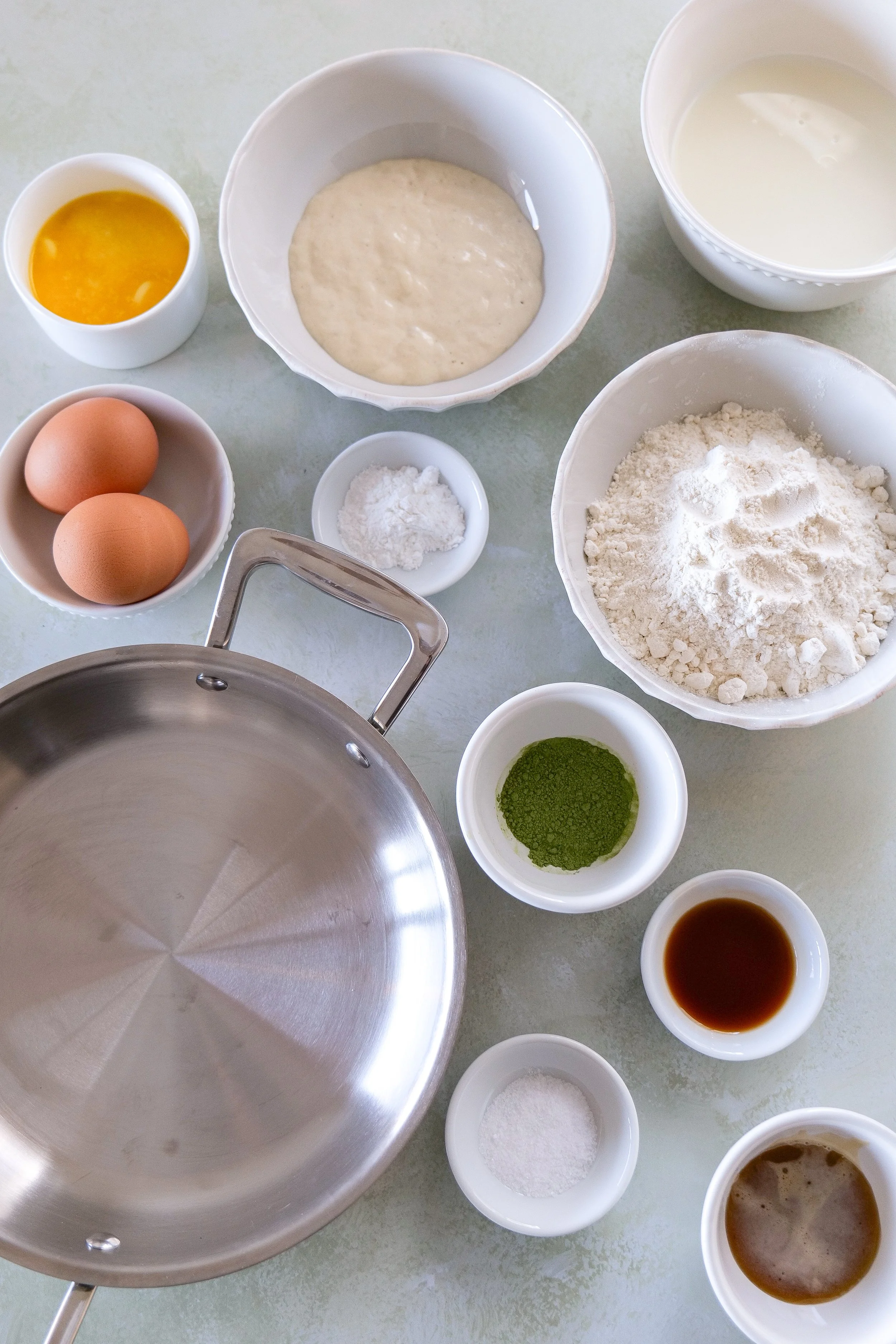 Ingredients for matcha sourdough discard pancakes including flour, matcha powder, eggs, milk, and sourdough discard on a kitchen counter