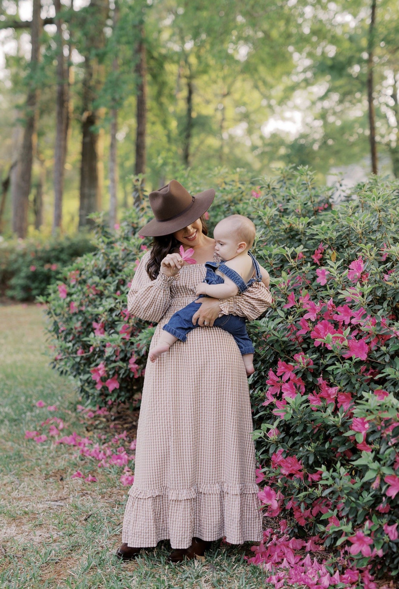 Mom holding her baby and showing him a pink flower beside blooming bushes at their Texas home