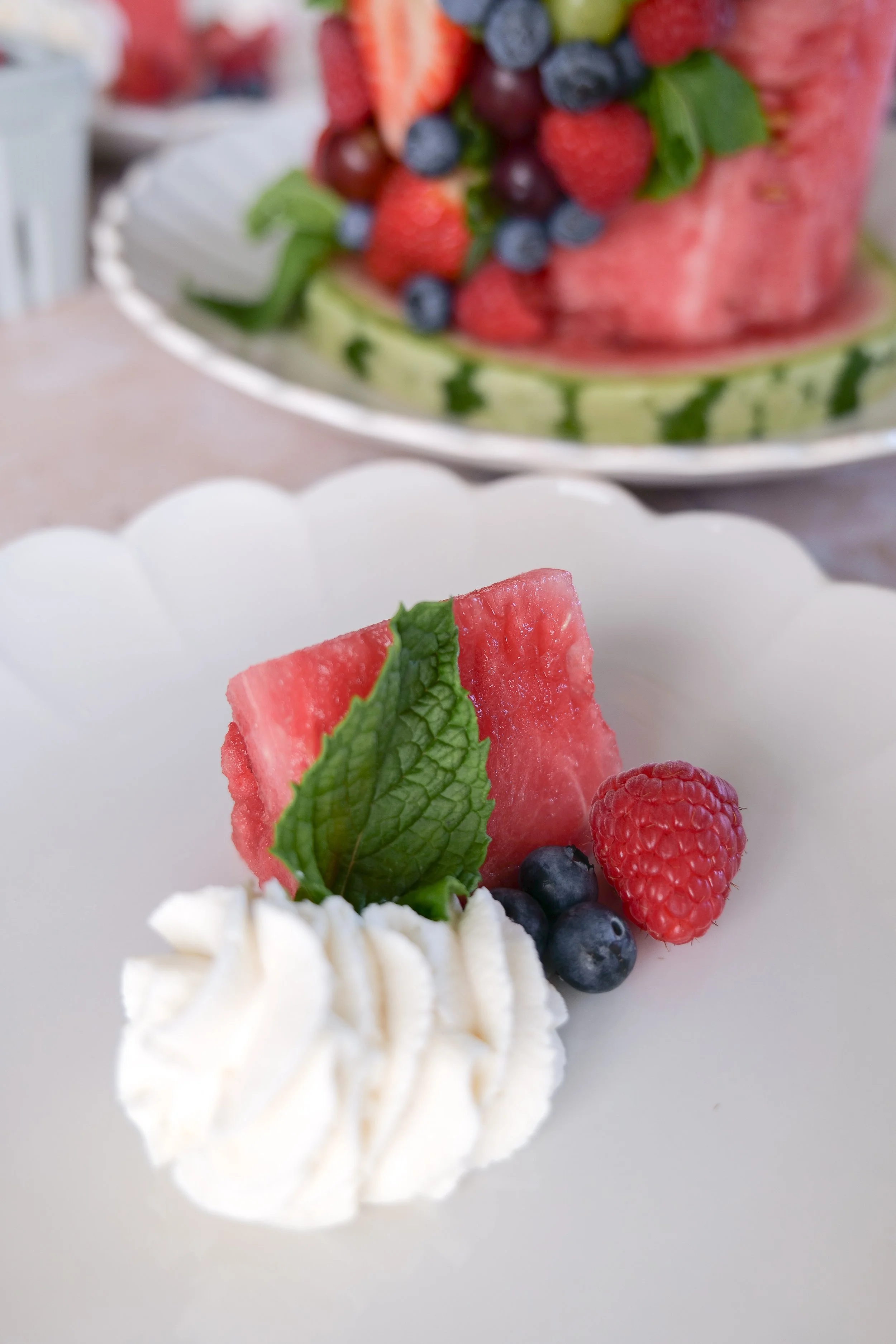 A slice of two-tier watermelon fruit cake served with homemade whipped cream on the side, fresh berries, grapes, strawberries, and mint for a refreshing summer dessert.