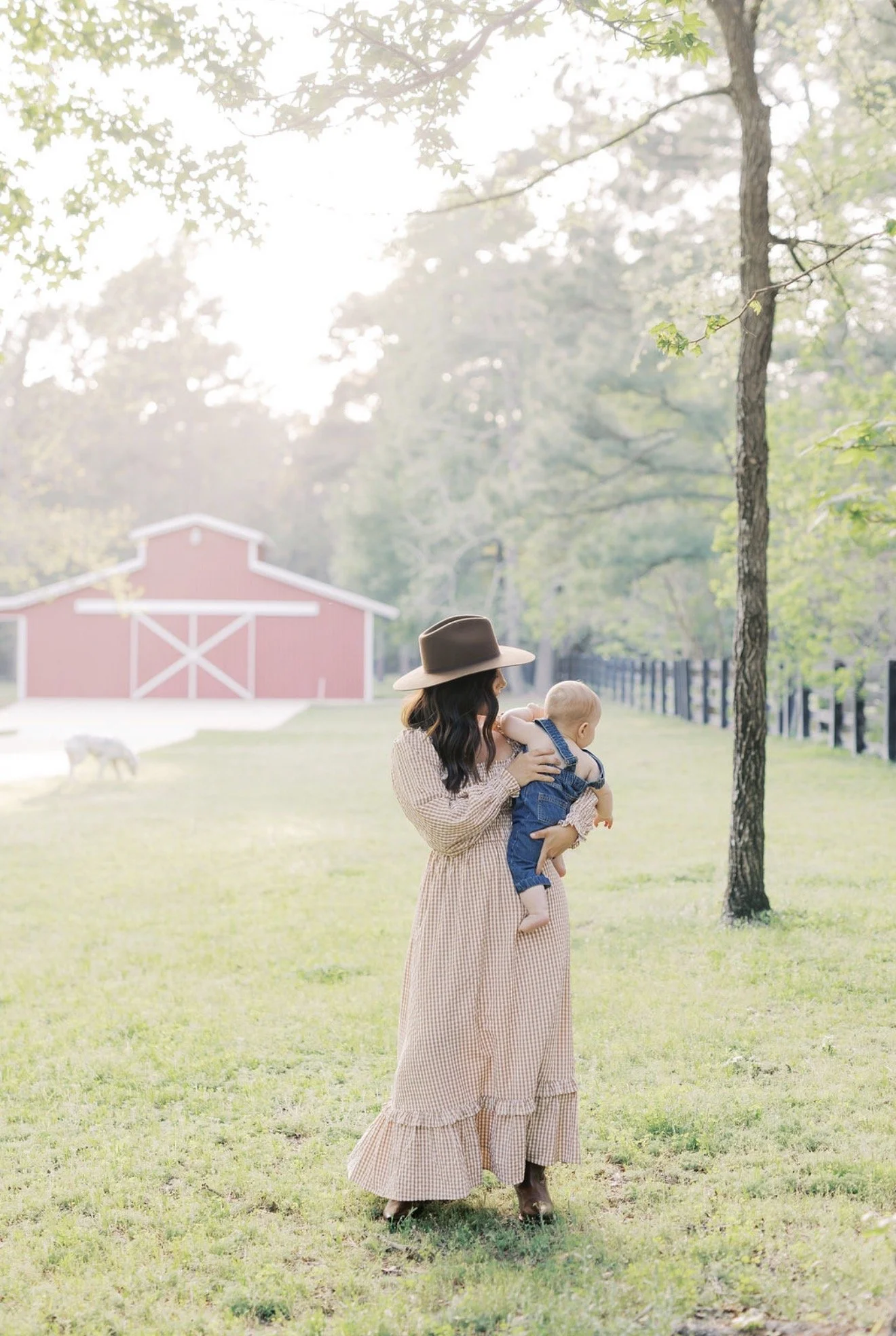 Mom in a cowboy hat holding her baby with a red barn and Dalmatian in the background on their Texas acreage