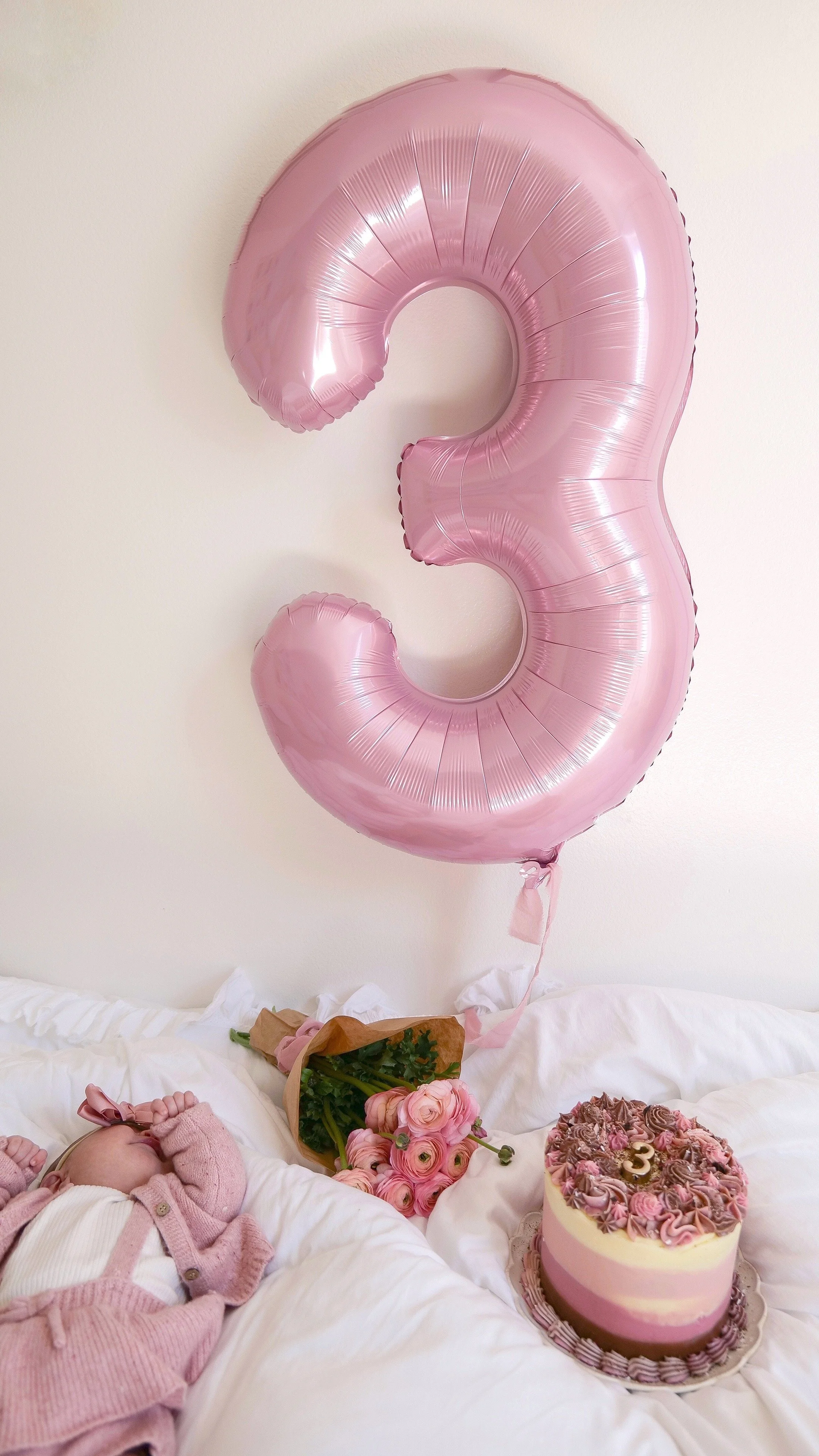 Neapolitan cake next to a bouquet of flowers and a number 3 balloon on a bed