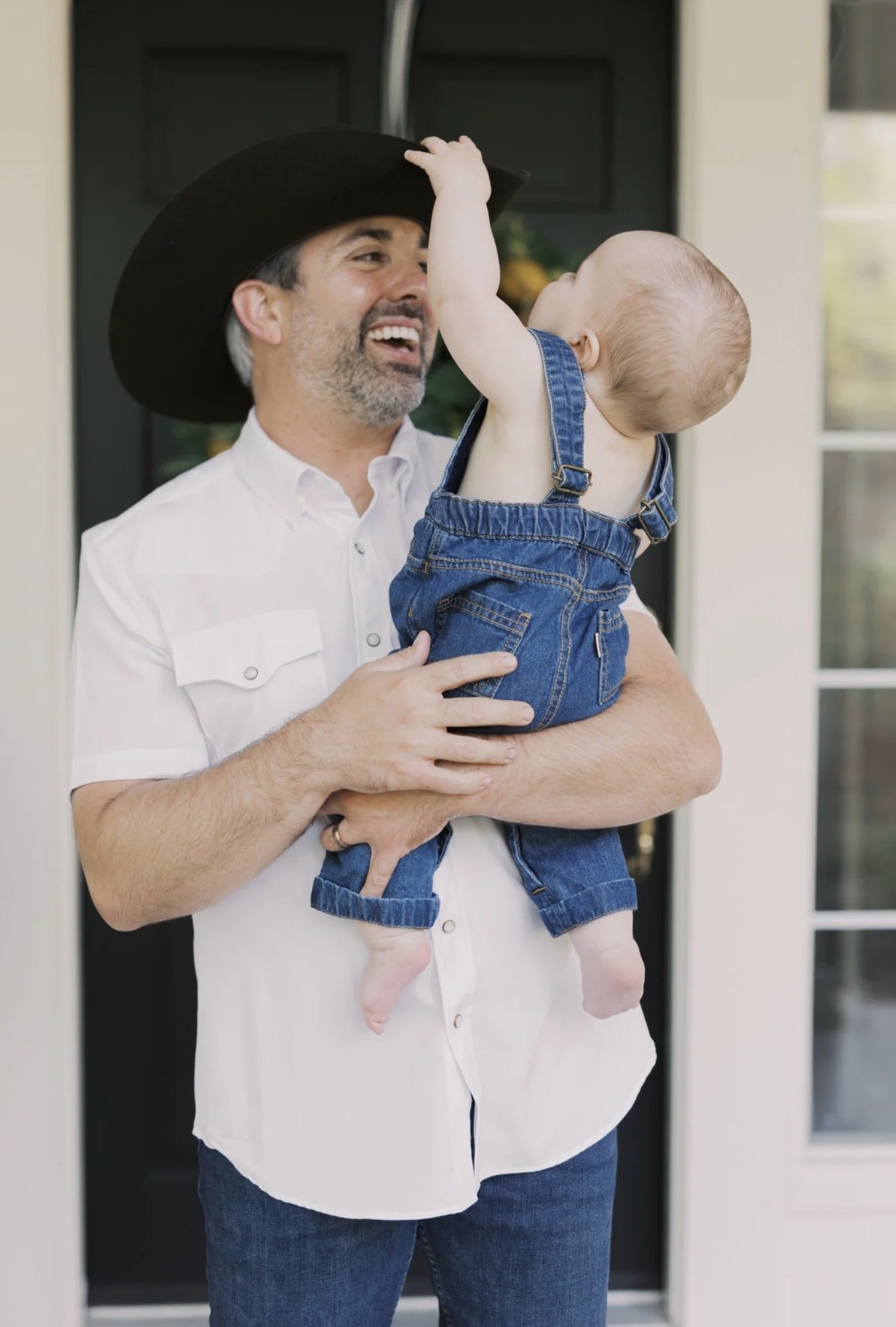 Dad holding his baby as he reaches for his cowboy hat during a family photo at their Texas home
