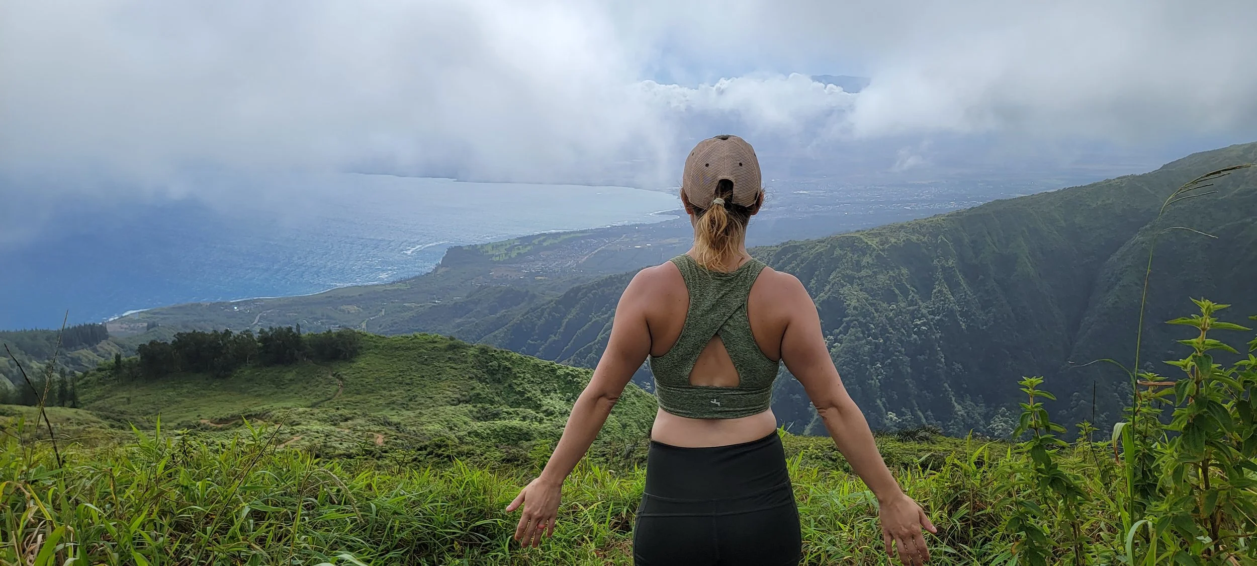 A woman stands outdoors with her arms slightly extended, facing a scenic view of green hills, a coastline, and a body of water under a cloudy sky in Maui, Hawaii. A photograph of author Cassandra Gelmanovich promoting her memoir Mother Maui.