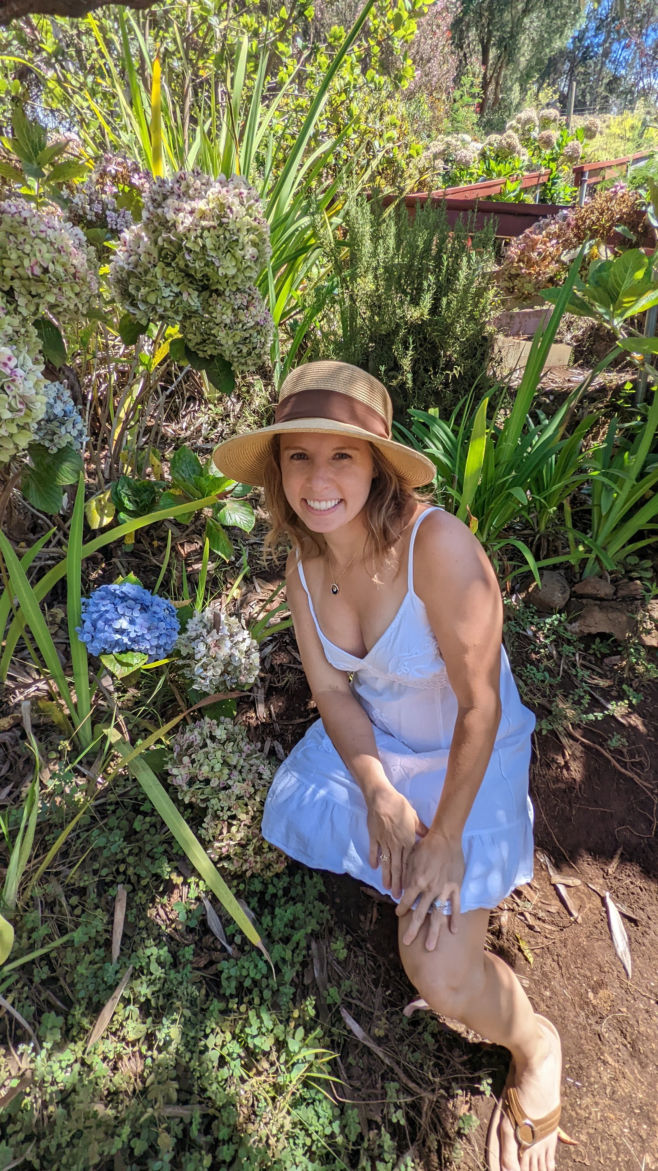 A woman in a white dress and wide-brimmed hat sitting in a garden among blooming hydrangeas and greenery. A photo of author Cassandra Gelmanovich in Maui, Hawaii showcased on her Sensitive Soul Press website.
