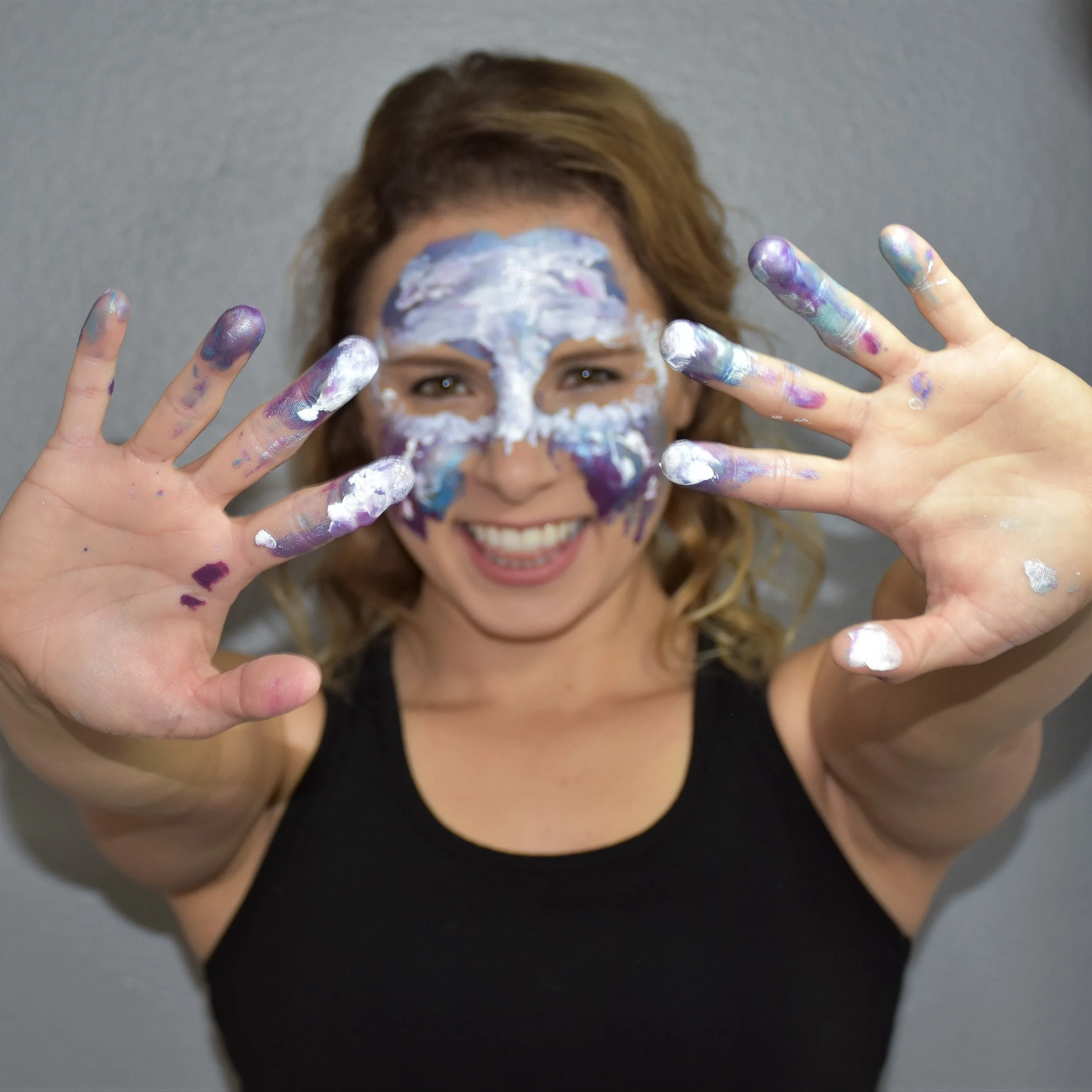 A smiling woman with face and hands covered in colorful paint, holding her hands up with fingers spread apart, against a plain gray background. An artistic bio photo of Energy Portrait artist Cassandra Gelmanovich.