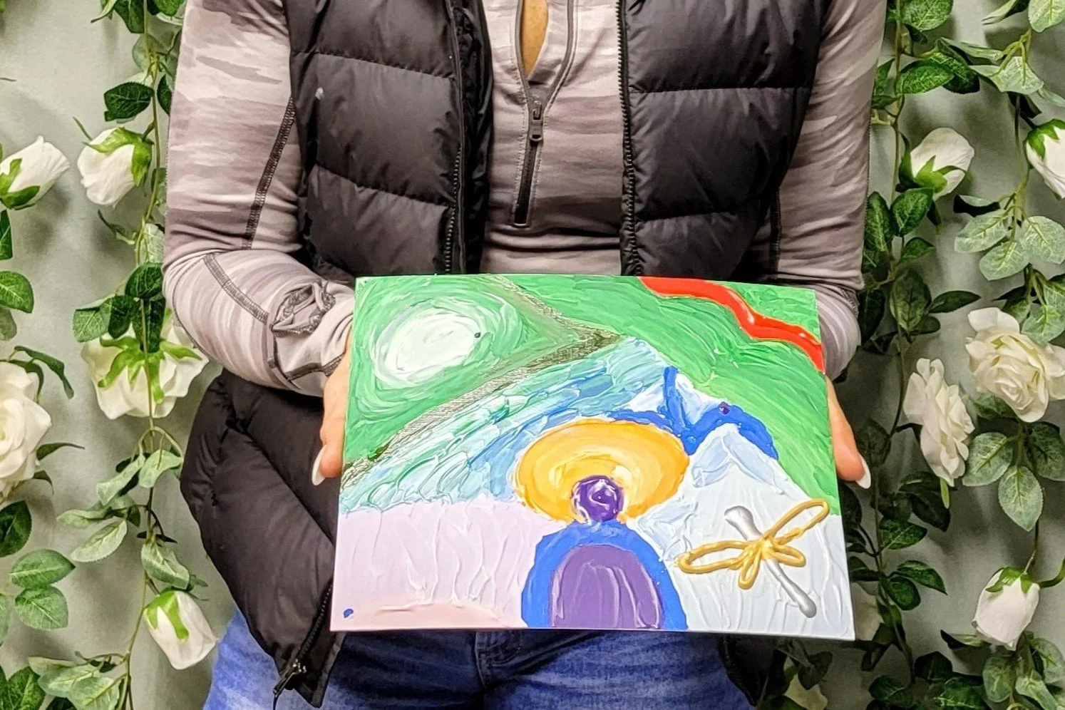 Person holding a colorful abstract painting, wearing a black vest over a gray long-sleeve shirt, standing in front of green leafy plants. An example of an Energy Portrait painted by artist Cassandra Gelmanovich with Sensitive Soul Fine Arts.