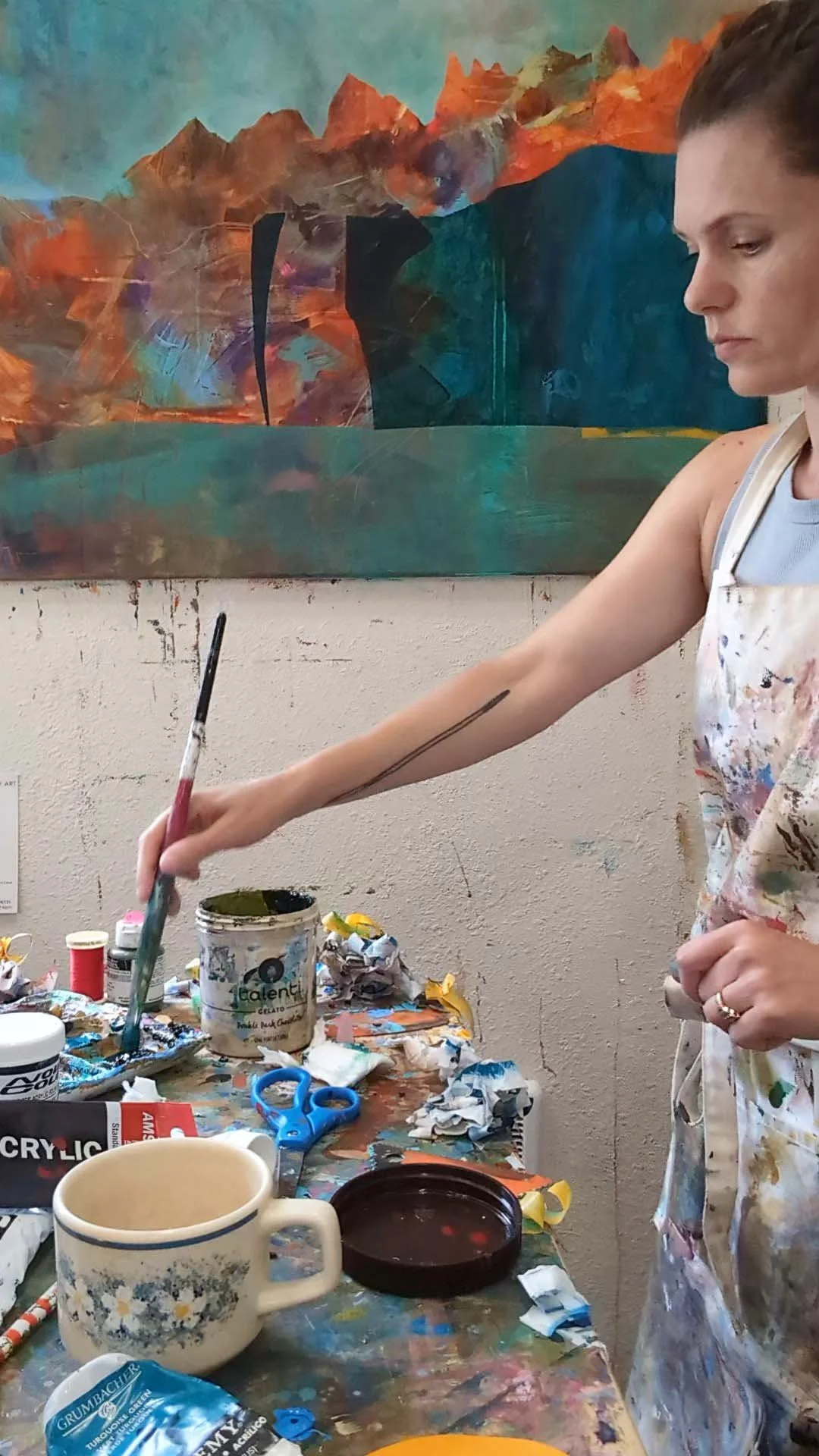Darby Guerechit, a landscape painter is standing at her work table in front of a piece in progress and dipping her paintbrush into a  paint pallette.