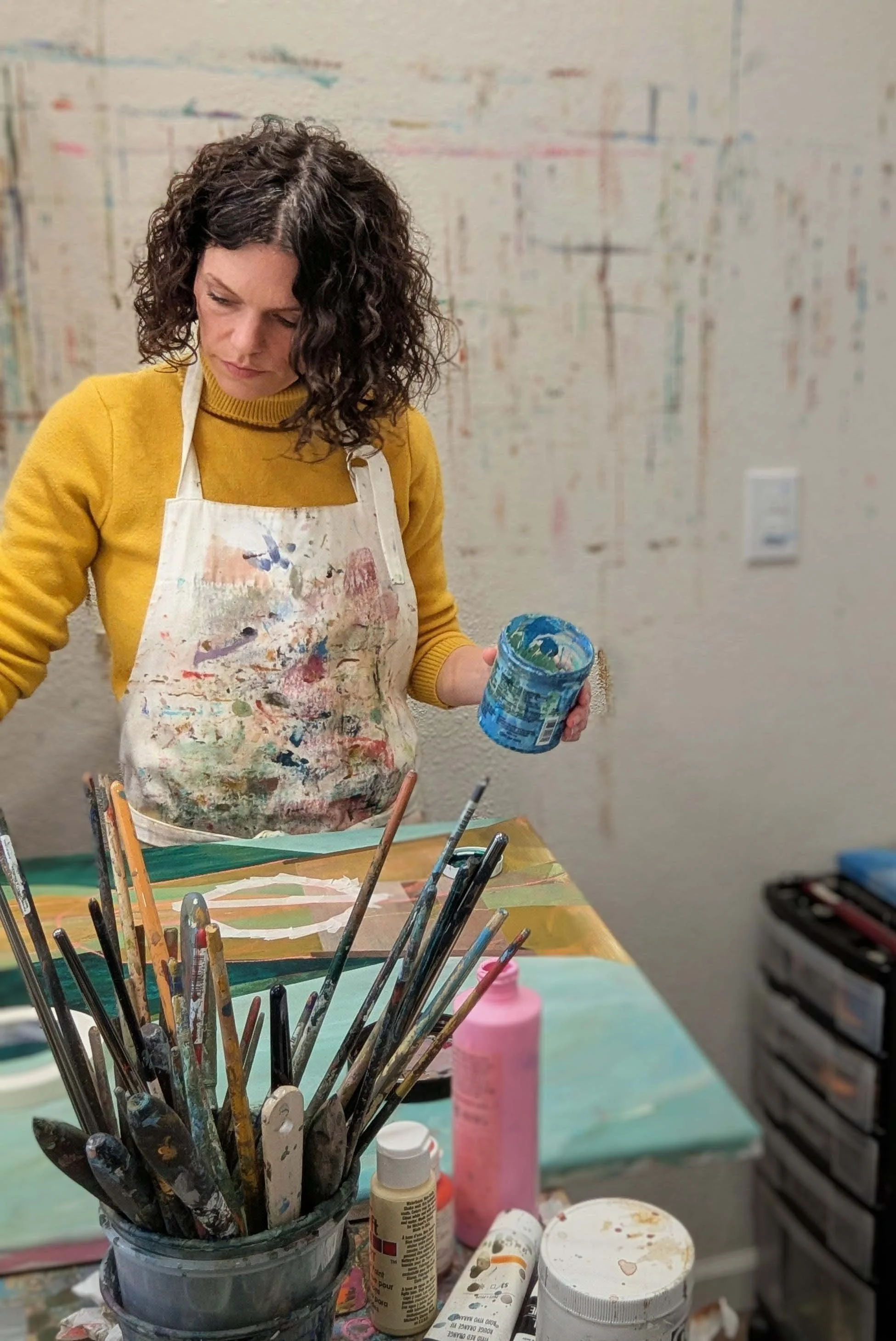 Darby Guerechit, landscape painter from the Pacific Northwest stands at her work table holding a cup of paint and painting on a canvas. She is wearing a yellow sweater and a painting apron.