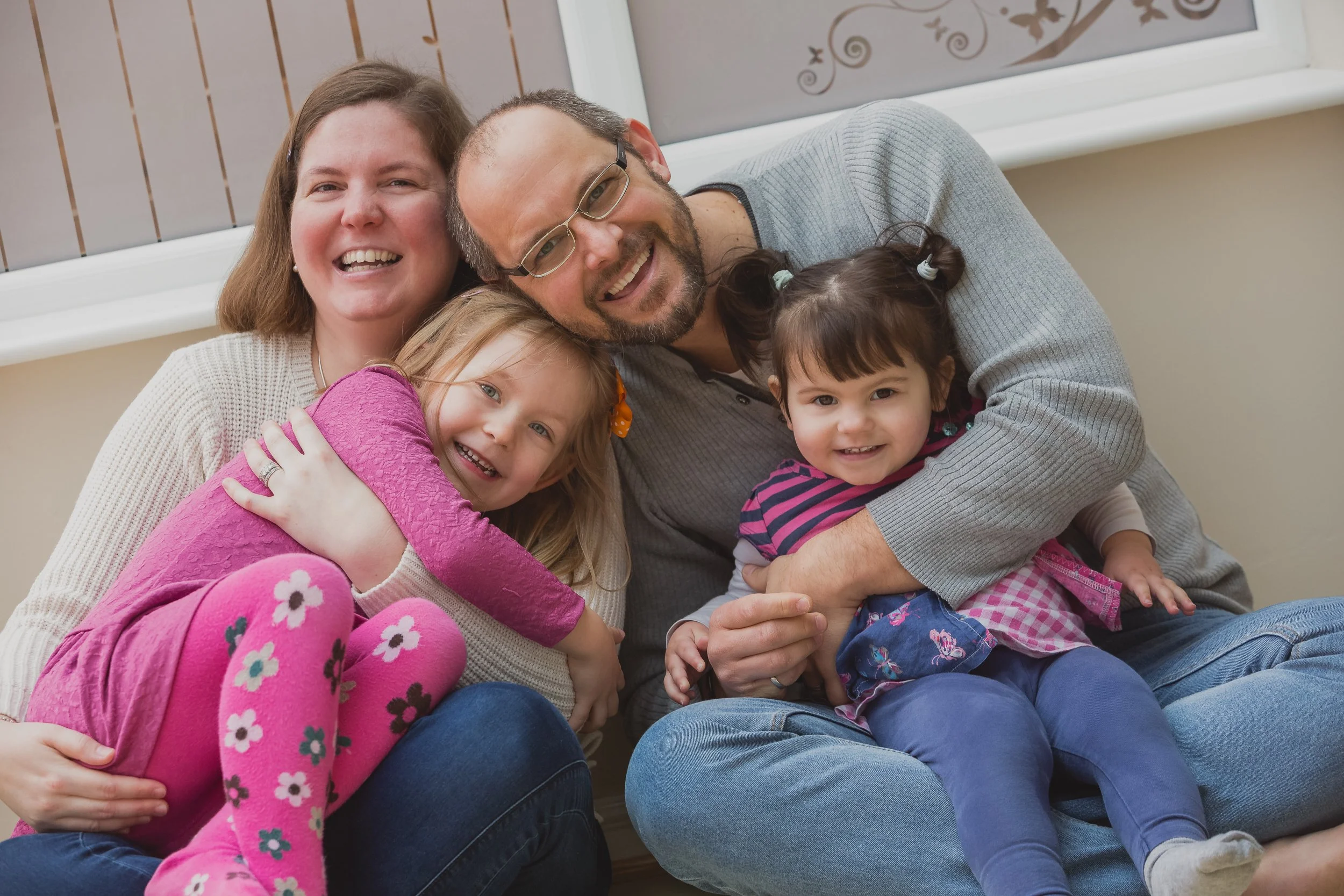 A cheerful family of four sitting close together indoors, smiling. The mother has shoulder-length brown hair, and the father has glasses and a beard. Two young girls are sitting with them, one with blonde hair in a pink dress and colorful leggings, and the other with dark hair in pigtails, wearing a striped shirt. The family is sitting on the floor near a window.
