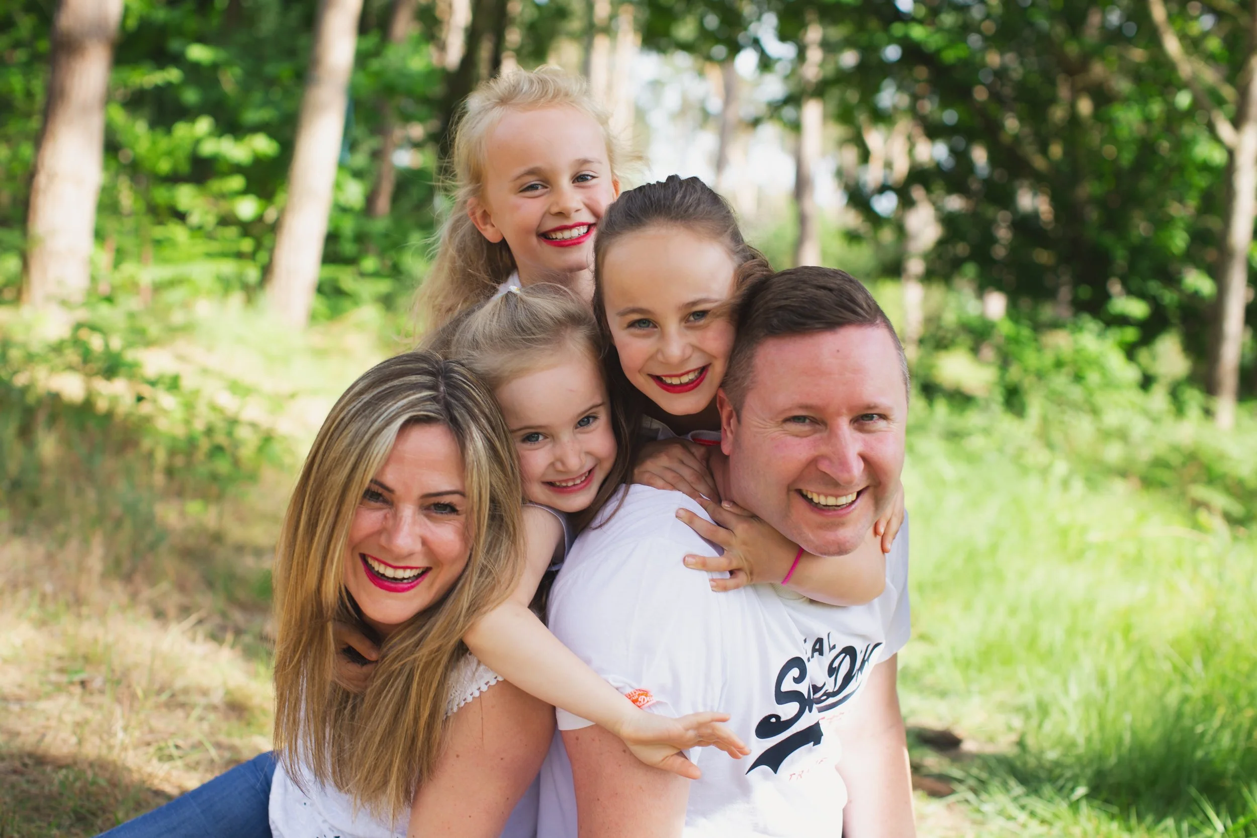 A happy family of six, including two women, one man, and three young girls, enjoying outdoors in a green forest setting.