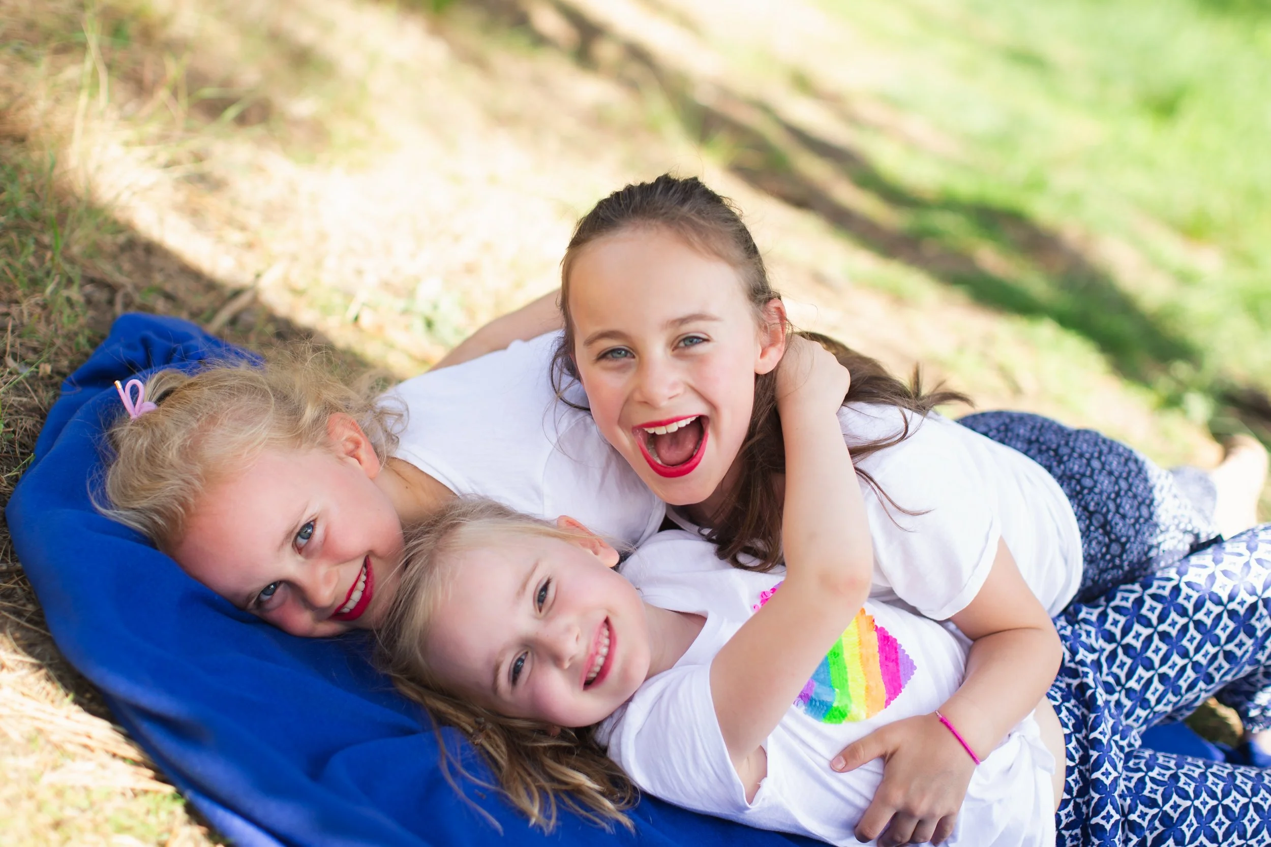 Three young girls lying on a blue blanket outdoors, smiling and laughing, with trees and grass in the background.