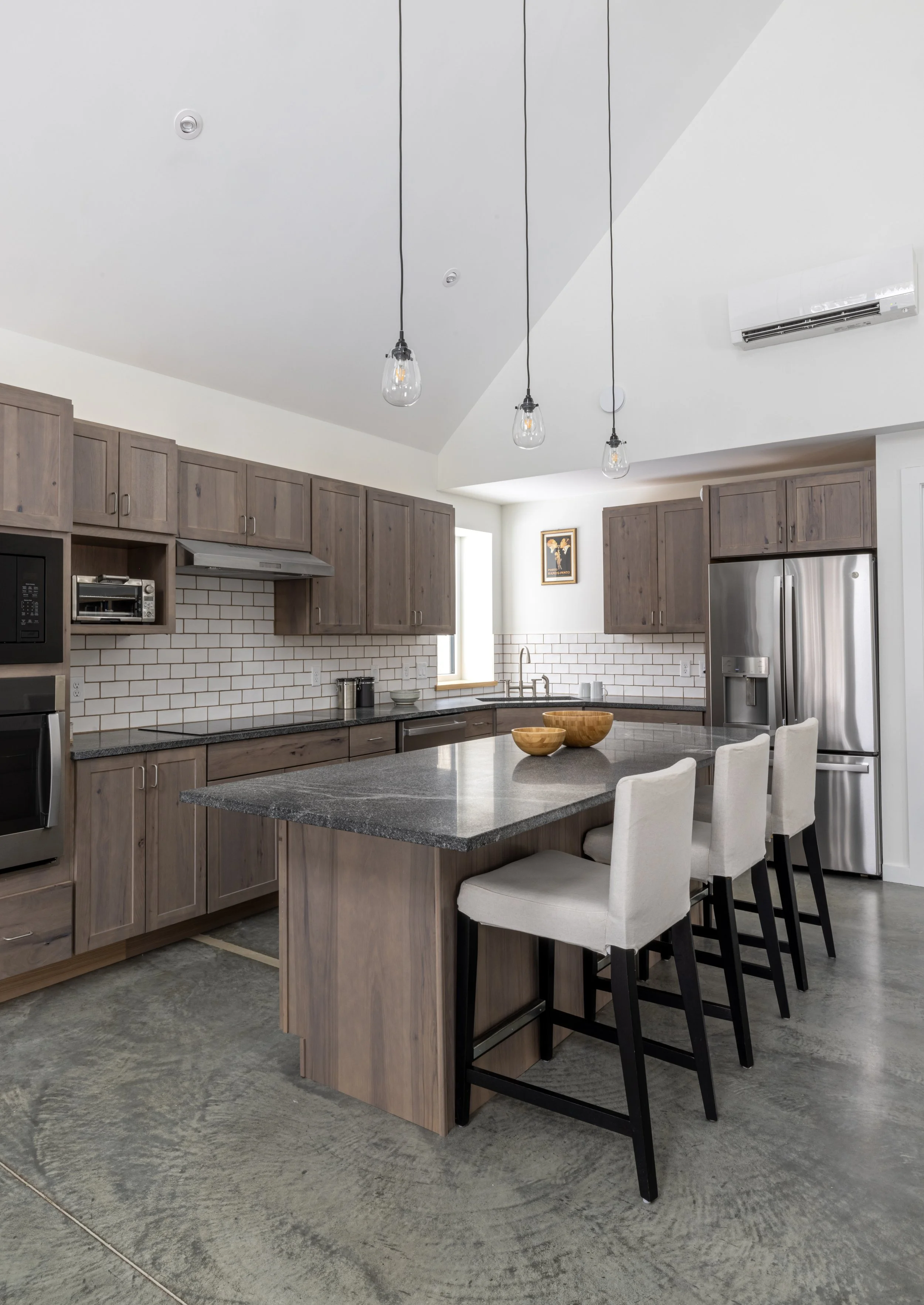 Kitchen with concrete floors, medium taupe cabinets, white subway tiles, and minimalist pendant lighting from the vaulted ceiling