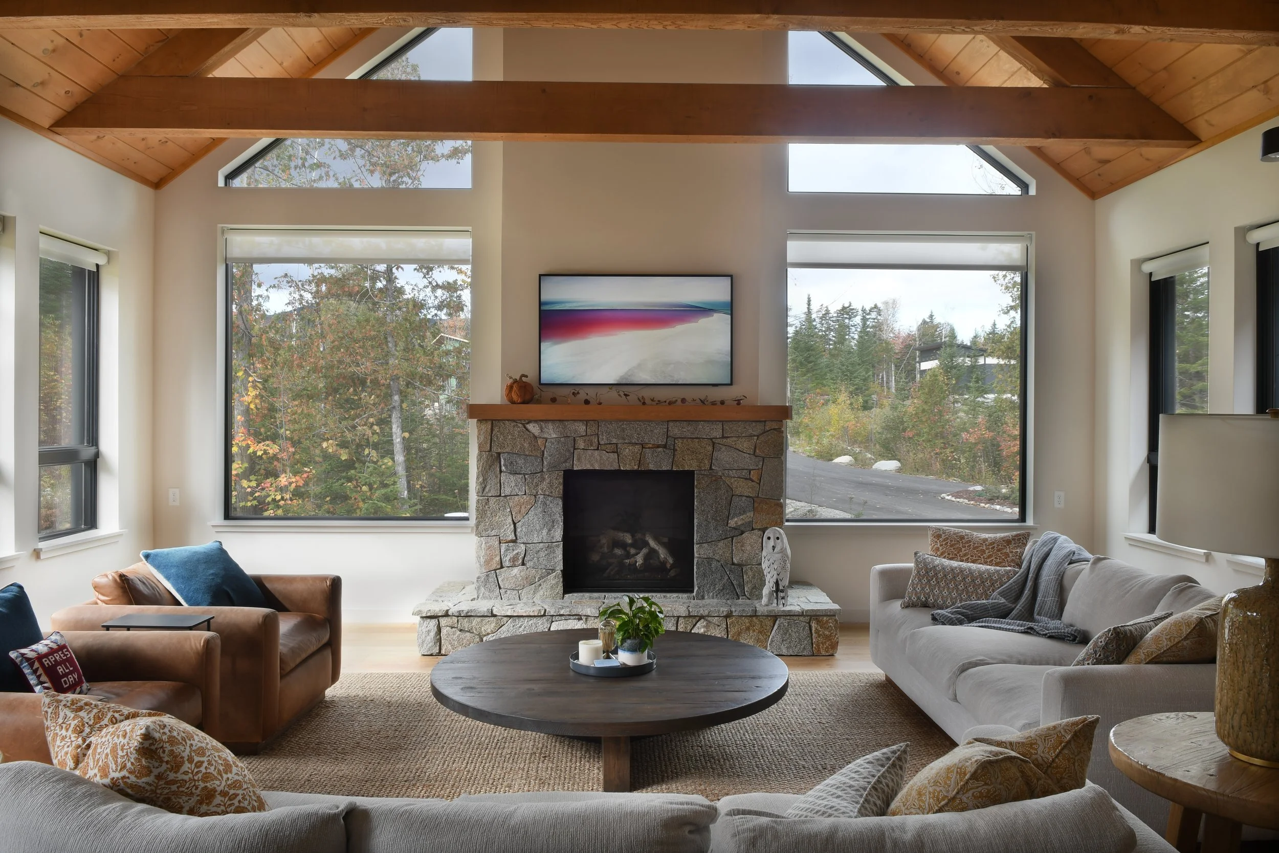 Inside the living room, couches and armchairs gather around a large circular coffee table and stone fireplace, with large windows showing the forest views.
