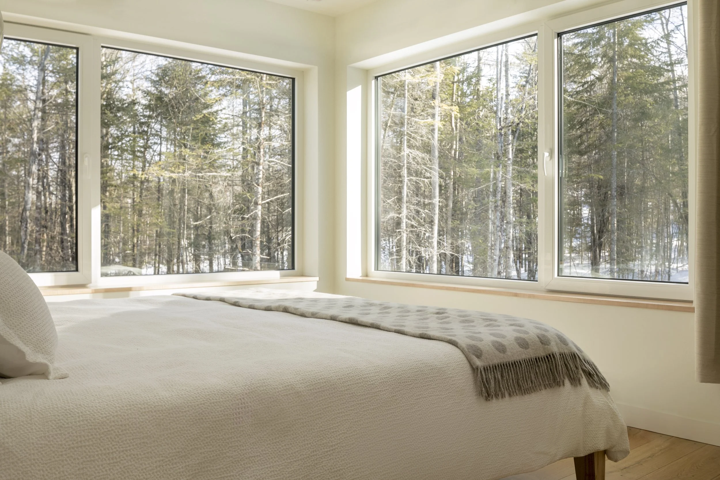 A bed with white and grey bedding faces two large windows with views of snowy pine trees