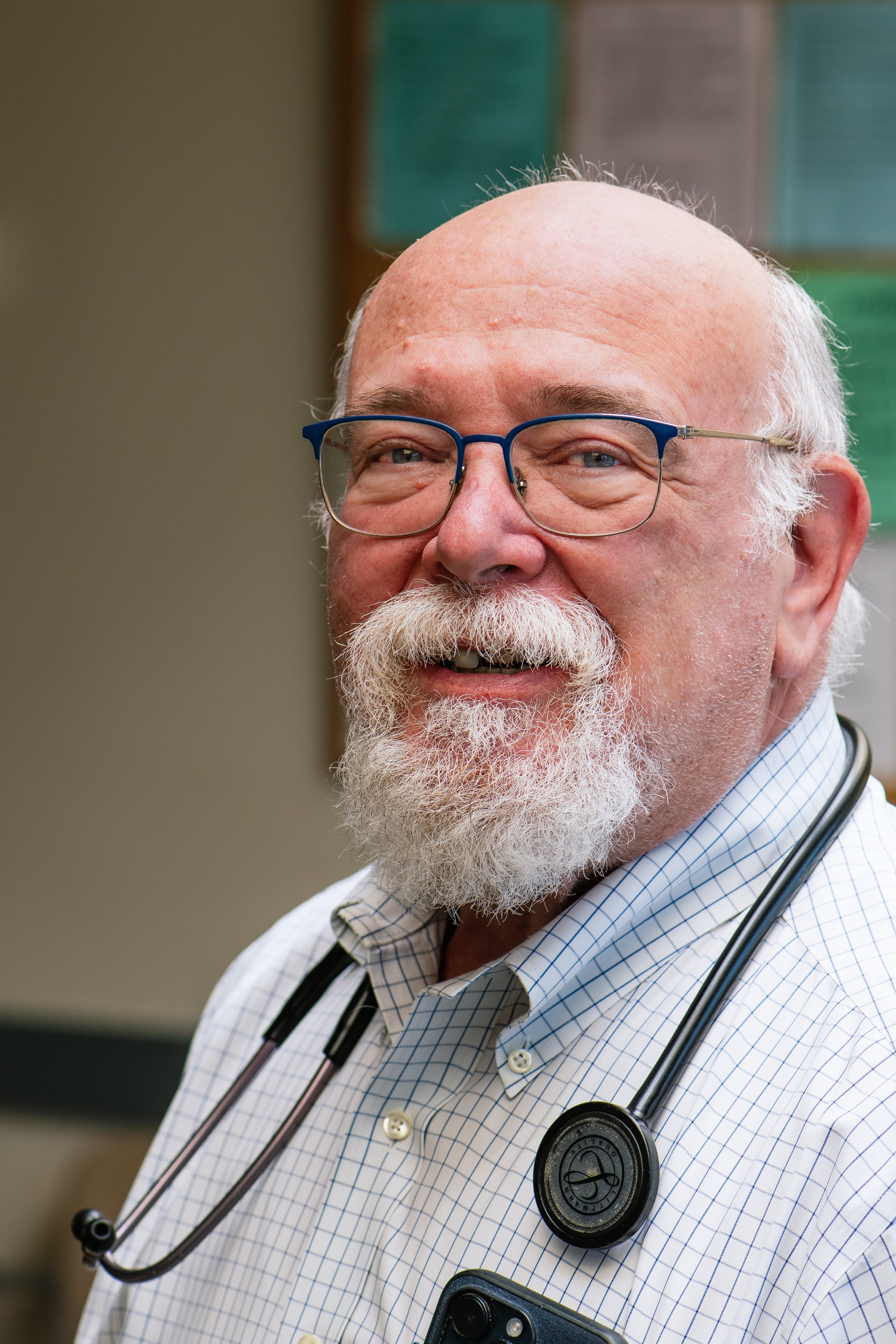 A smiling elderly male doctor with glasses, a white beard, wearing a checked shirt and a stethoscope around his neck, in a medical office.