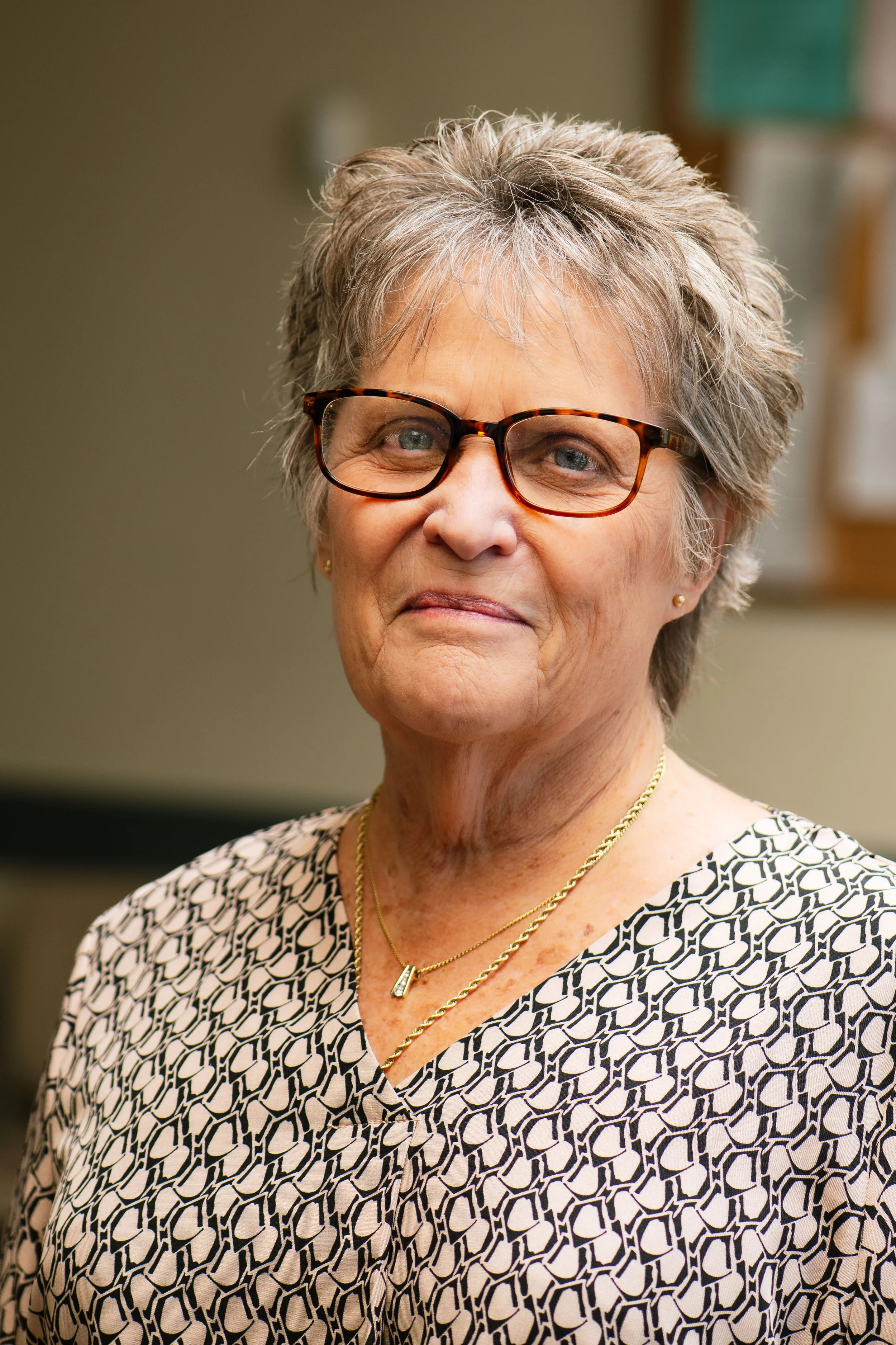 A portrait of an older woman with short gray hair and glasses, wearing a patterned blouse and gold jewelry, smiling softly in an indoor setting.