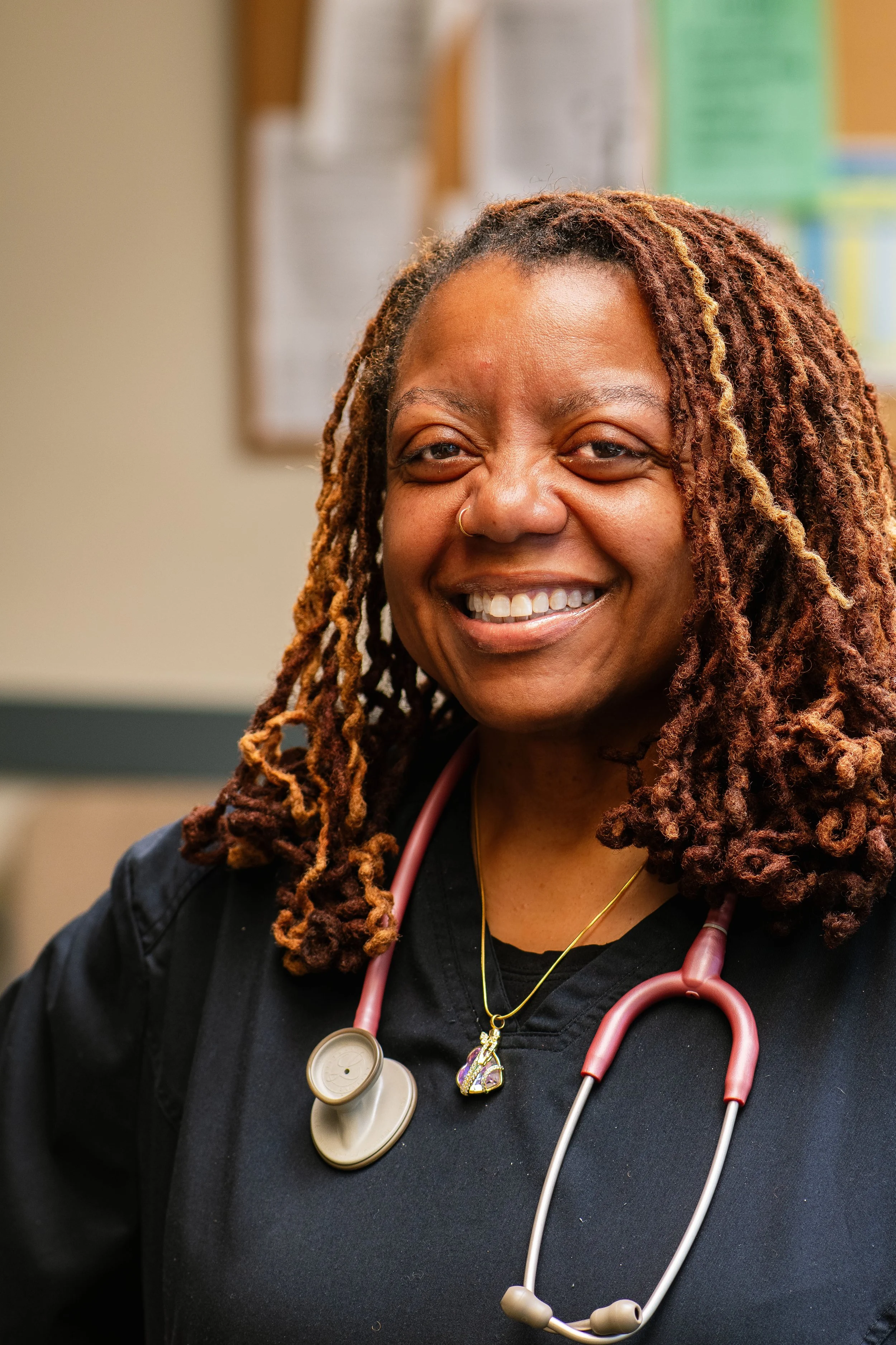 A smiling female healthcare professional with dreadlocks, wearing a black uniform and a stethoscope around her neck, standing in a medical setting.