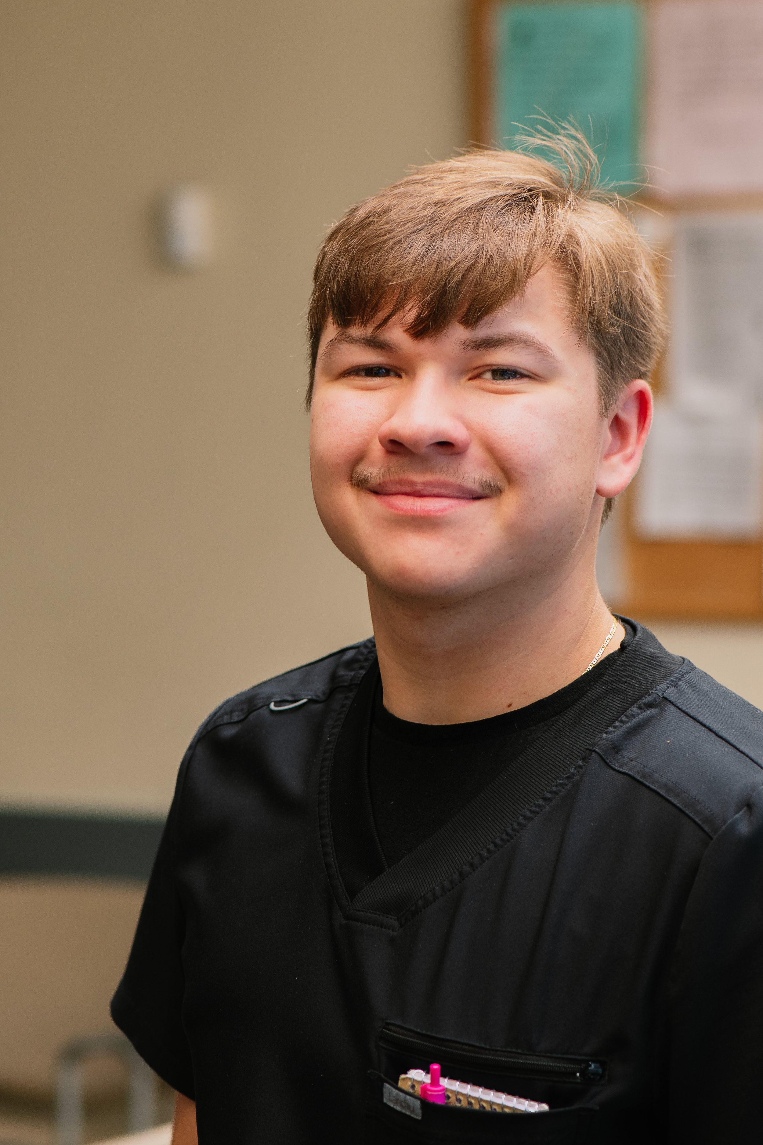 A young man in a black medical uniform smiling indoors with a bulletin board in the background.