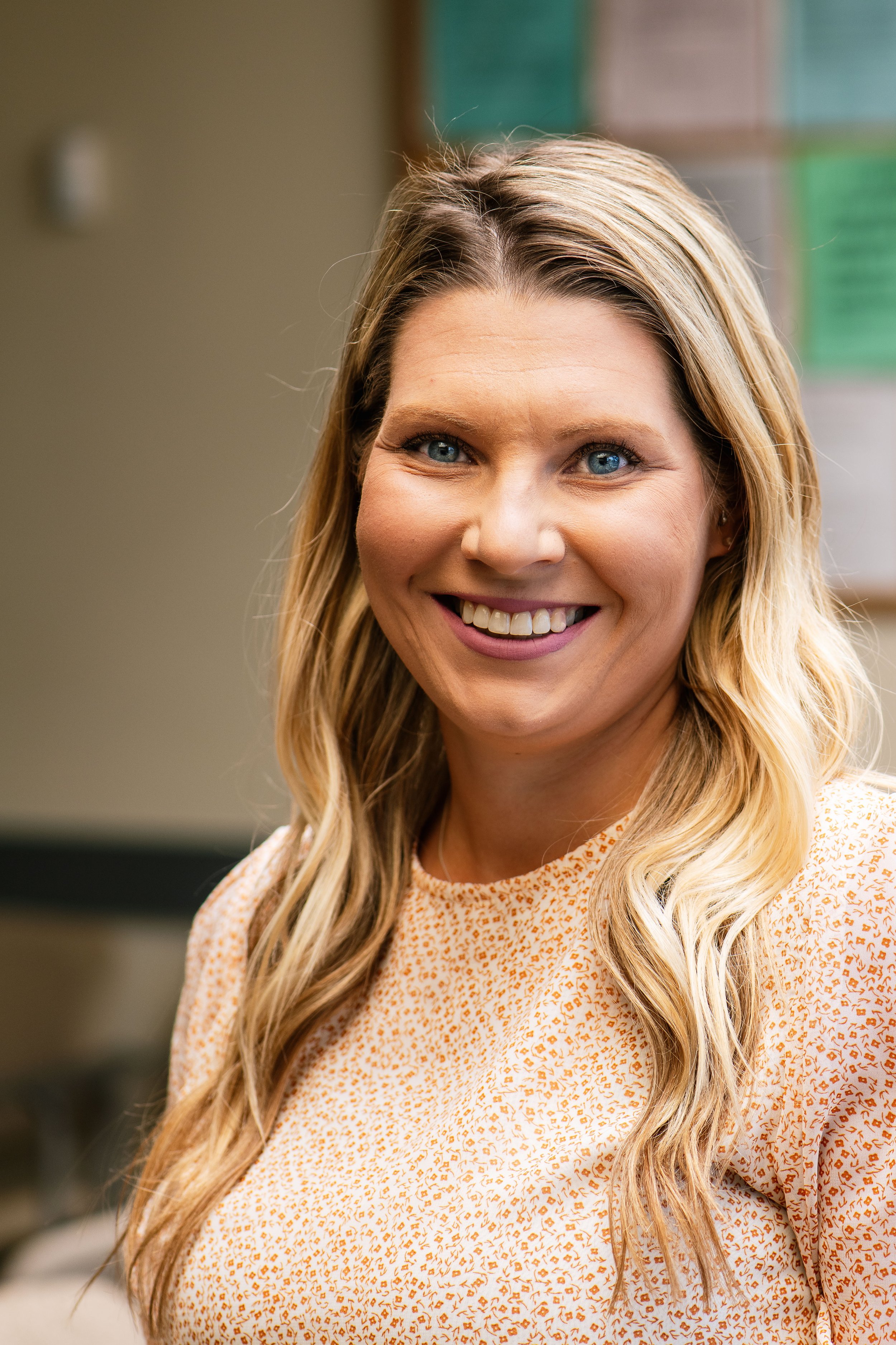 A smiling woman with long blonde hair, wearing a white and orange patterned top, in an indoor setting with erasers and papers on the wall in the background.