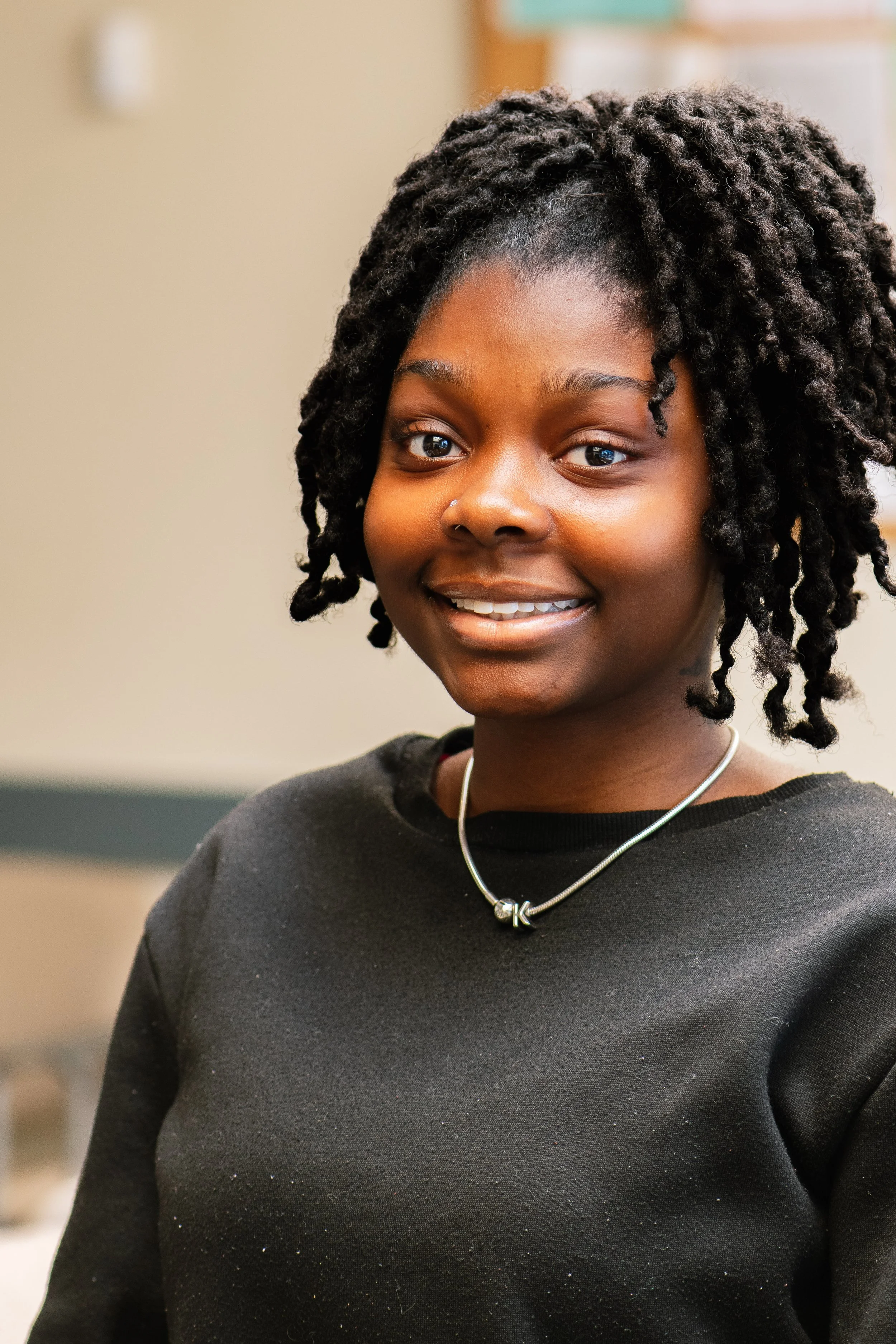 A young woman with short, curly hair and dark skin, smiling and wearing a black shirt and a silver necklace, indoors.