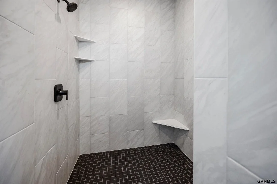 Empty shower with white tiled walls, black shower fixtures, corner shelves, and a dark tiled floor.