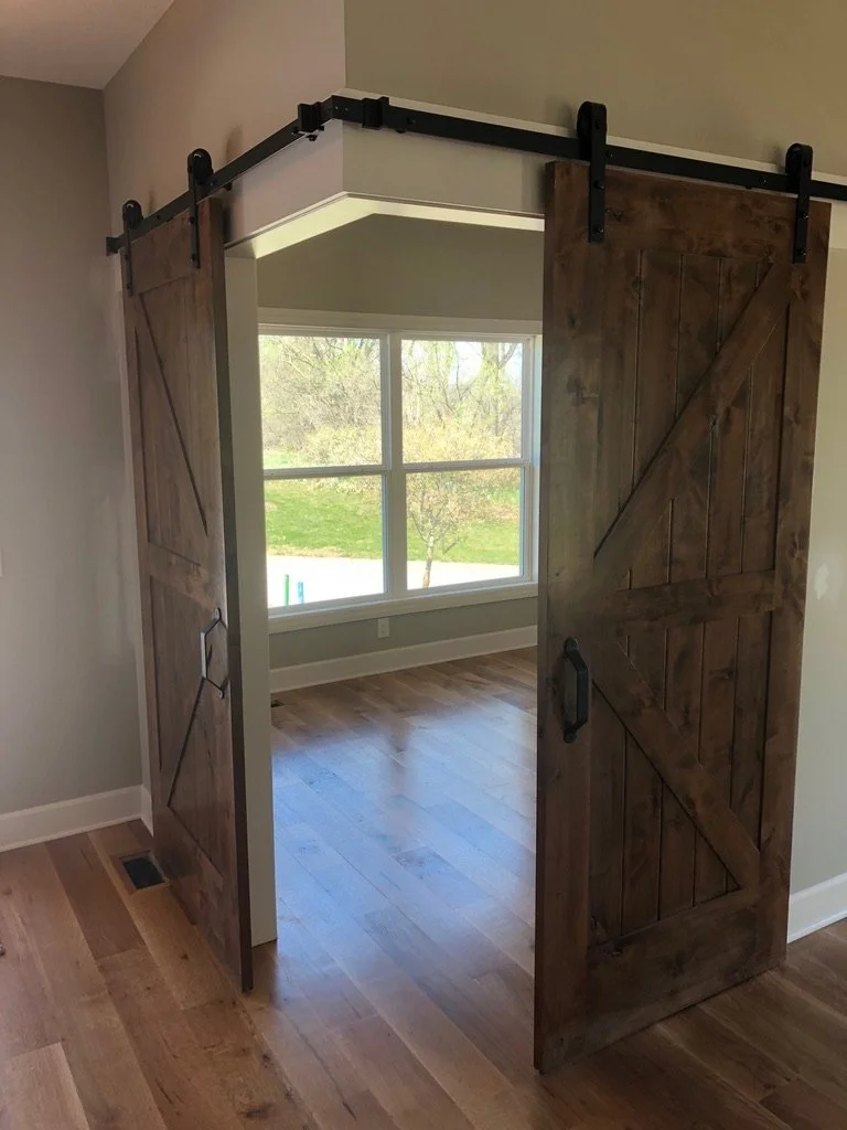 A pair of rustic wooden sliding barn doors on a black metal track in a house interior, opening to a room with a large window showing a view of a grassy outdoor area with trees.