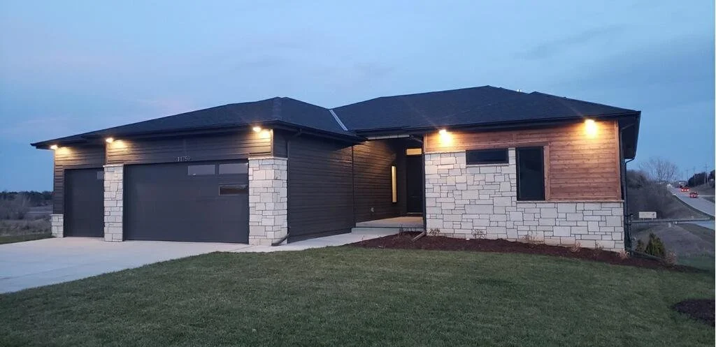 Modern house with stone and wooden exterior, illuminated by exterior lights at dusk, with a garage and a well-maintained lawn.