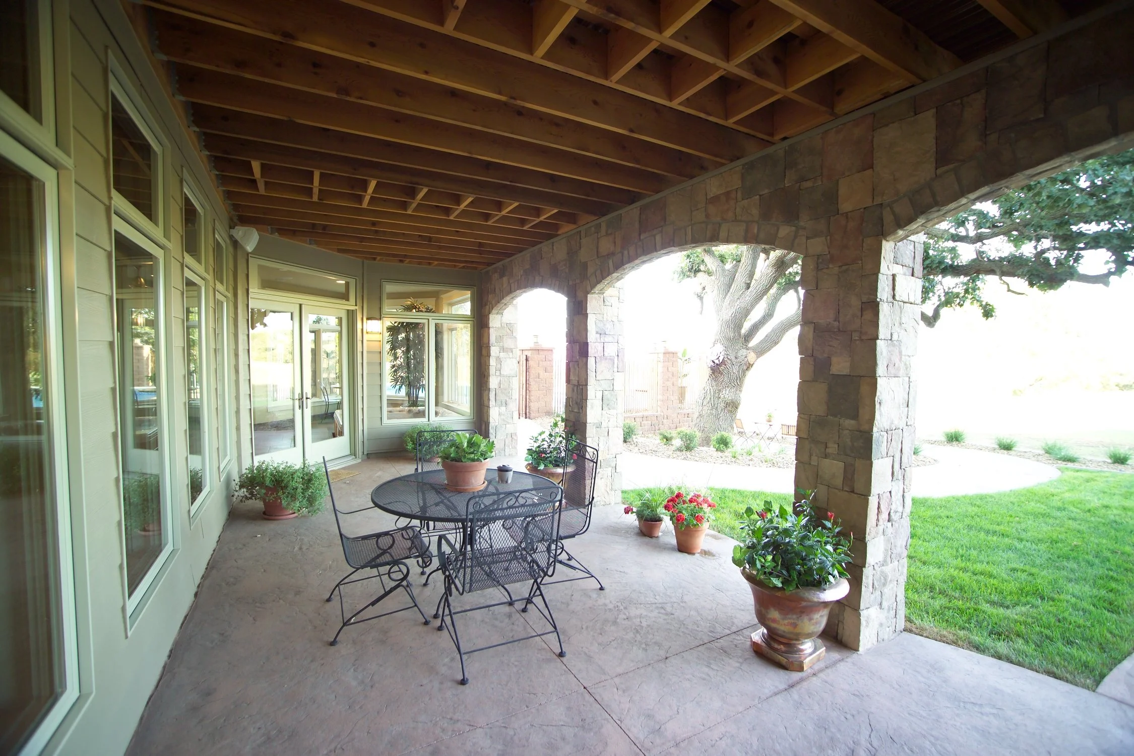 Covered patio with metal table and six chairs, potted plants, arched stone columns, and a view of a grassy yard with a large tree.