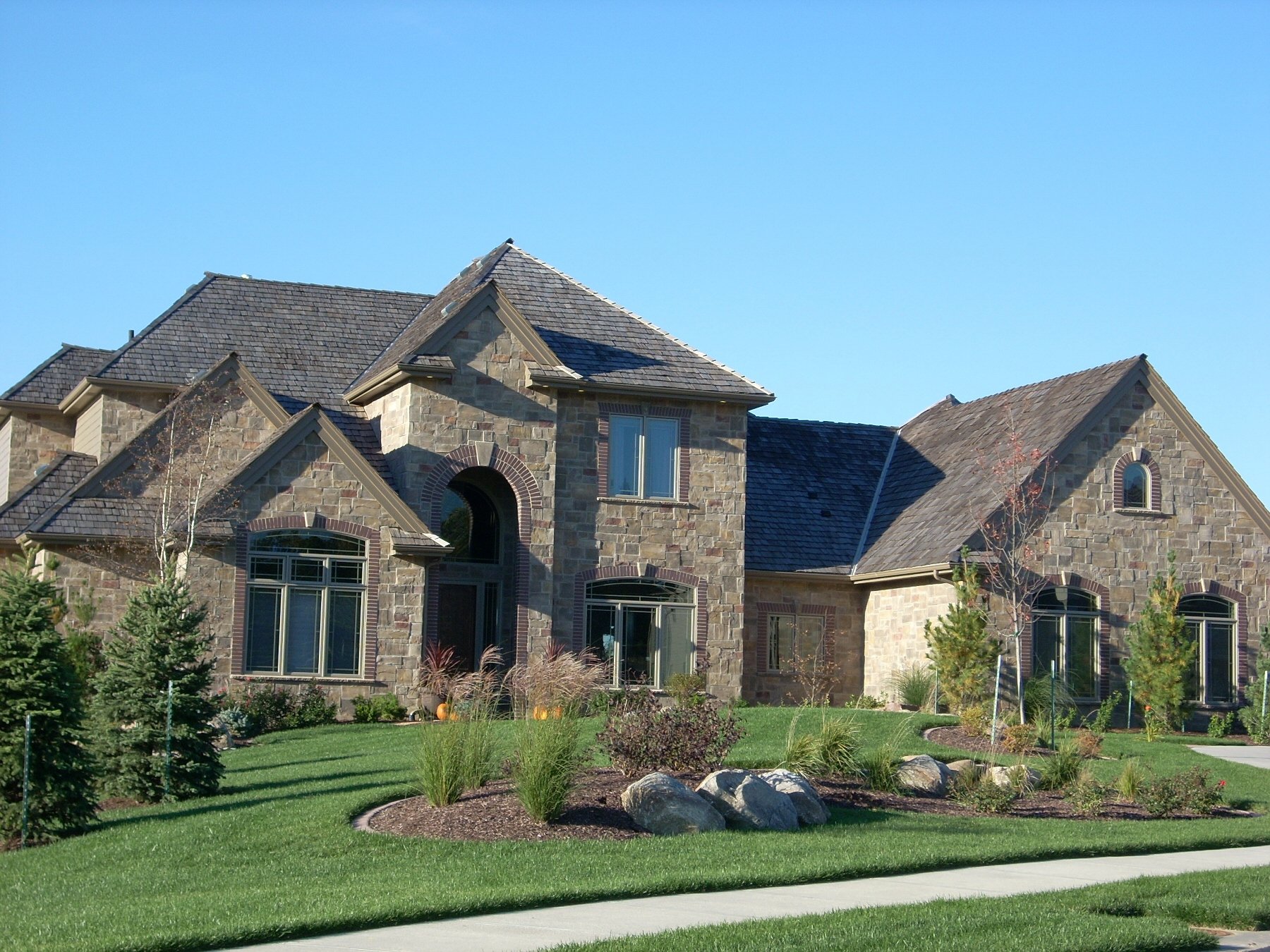 Large stone house with multiple pointed roofs, landscaped front yard with grass, rocks, small trees, and shrubs, under clear blue sky.