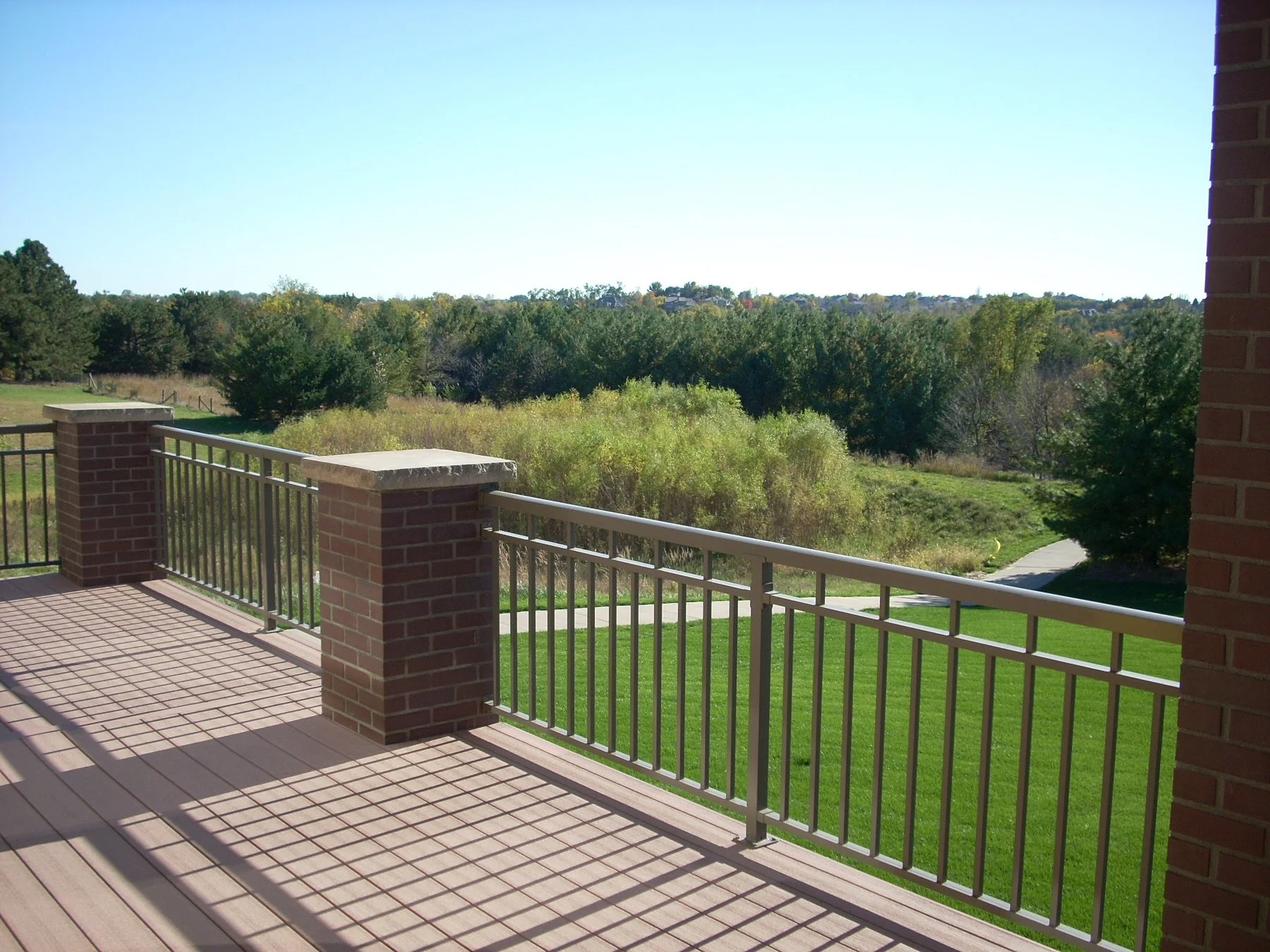 A view from a brick balcony with metal railing overlooking a grassy yard and trees under a clear blue sky.