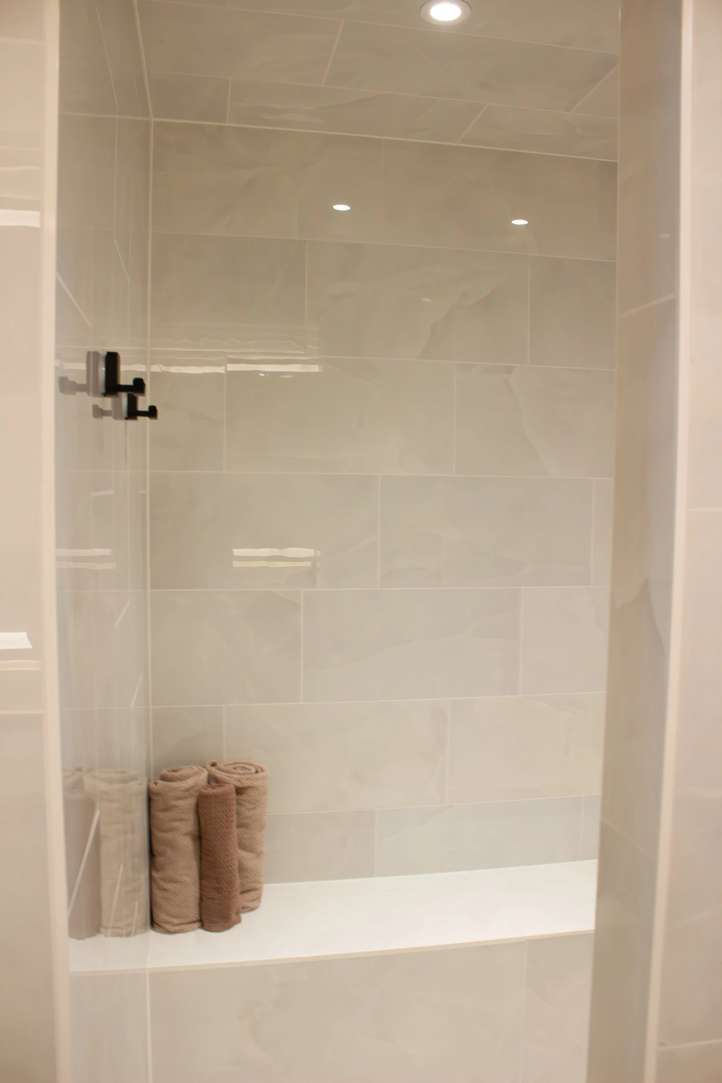 Empty walk-in shower with beige tiles, black hooks on the wall, and three rolled-up towels on a white bench inside the shower.