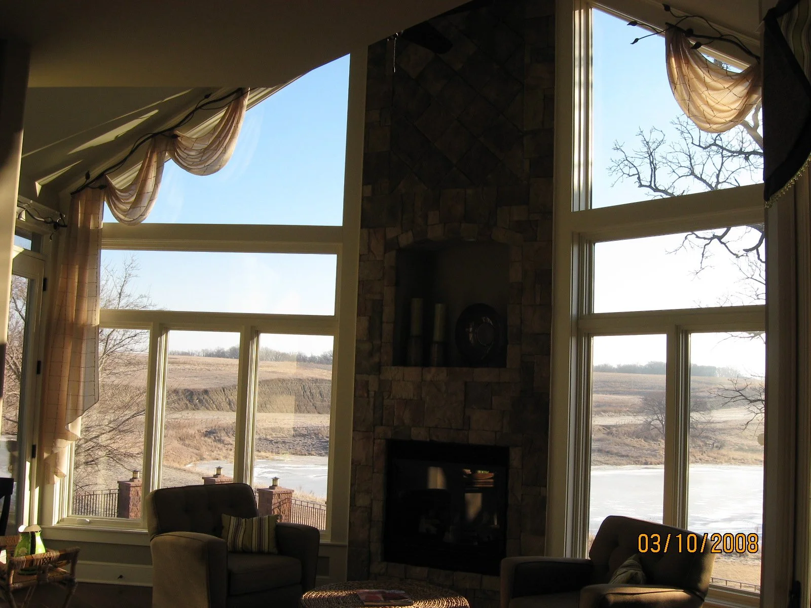 Living room with large windows showing a lake and hillside outside, stone fireplace, armchairs, and curtains.