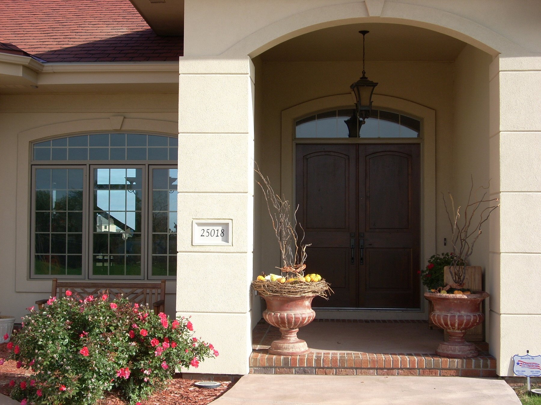 Front entrance of a house with large wooden double doors, a hanging lantern above the door, flower pots with bare branches, and a bush with pink flowers.