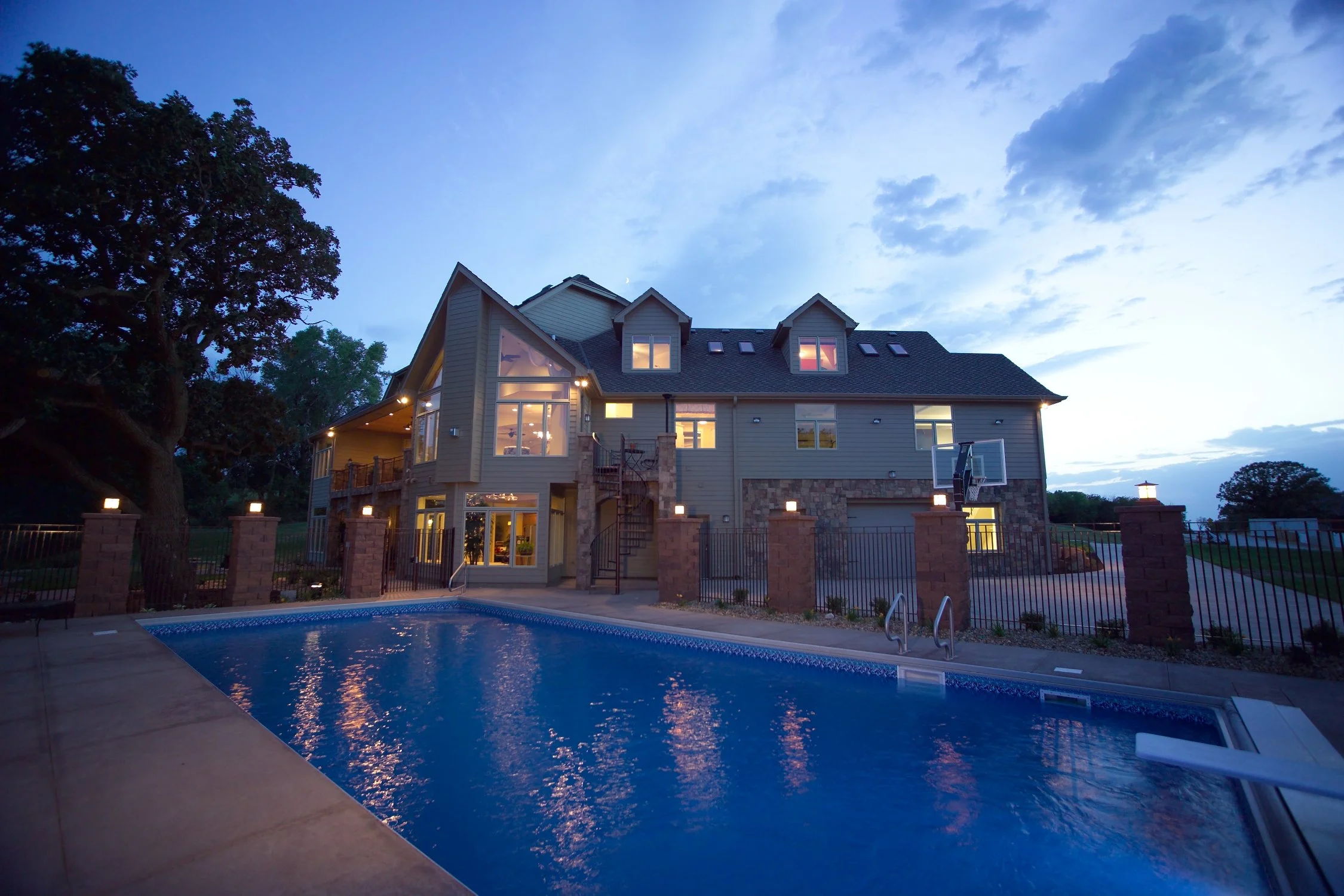Large modern house with lit windows, a swimming pool in the foreground, and a partly cloudy evening sky.