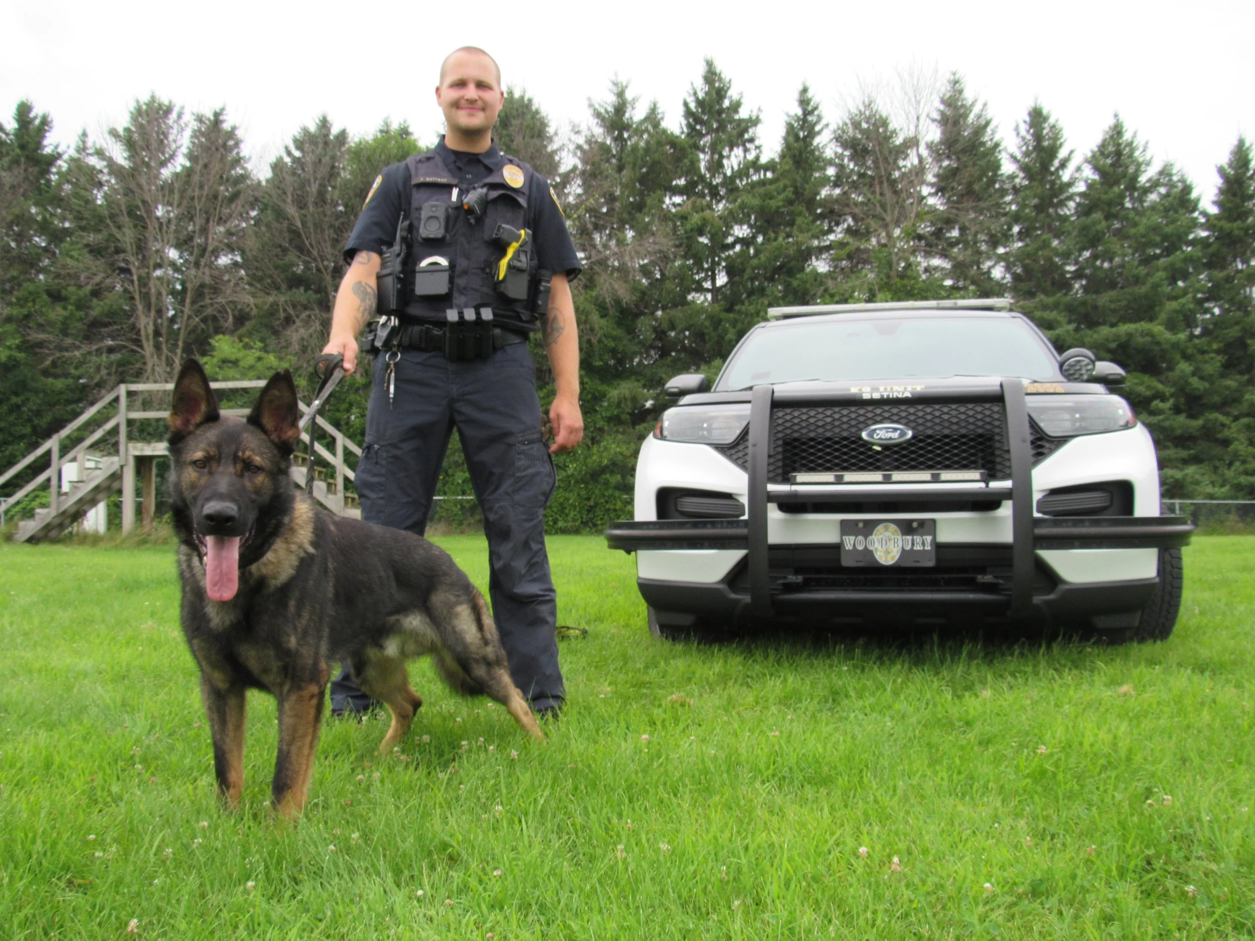 A police officer standing with a German Shepherd dog in front of a police car, with trees in the background.