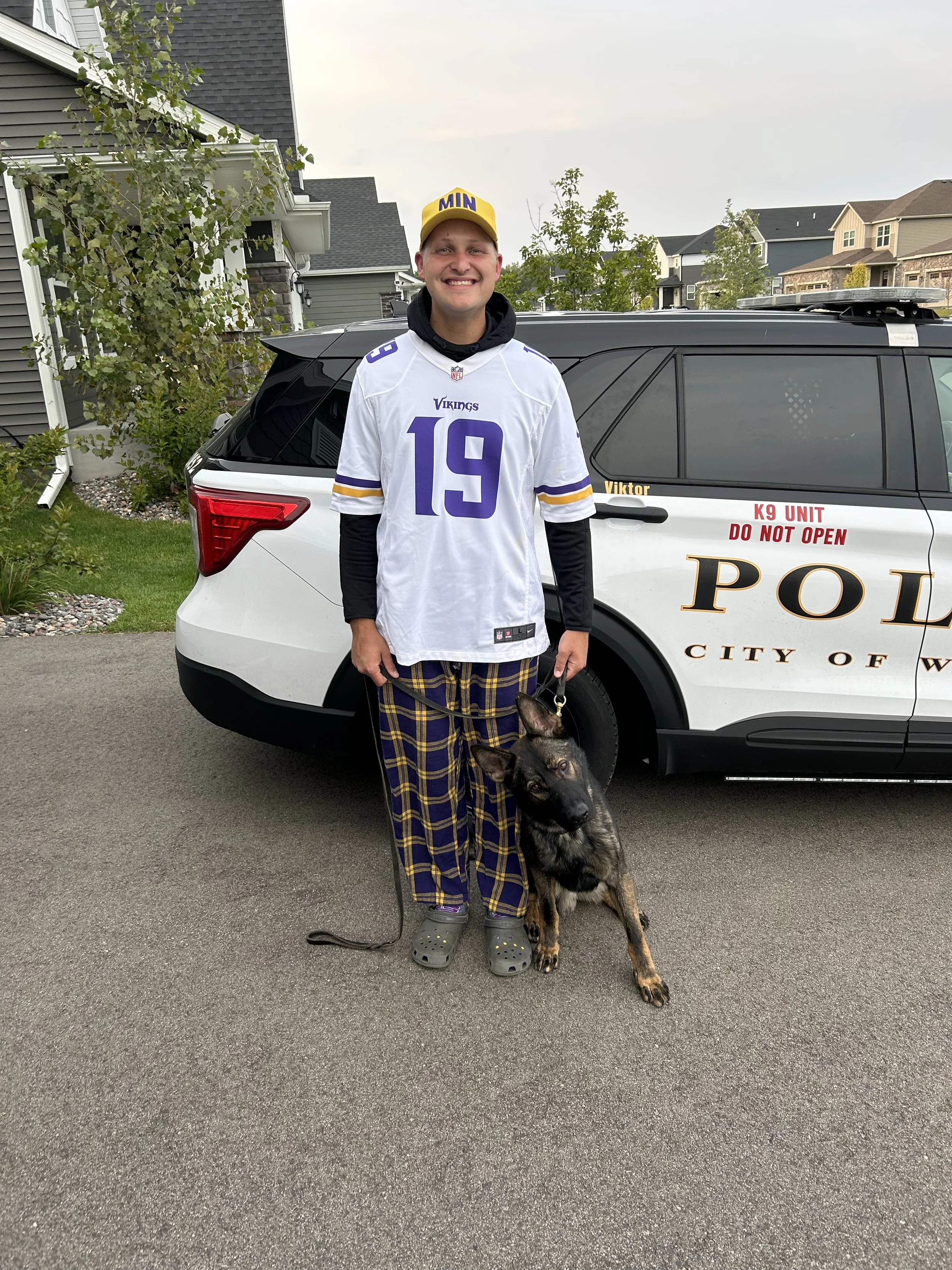 A young man standing outdoors on a gray asphalt driveway, smiling and holding a leash attached to a German Shepherd dog. He is wearing a white football jersey with purple and yellow accents, plaid pajama pants, gray Crocs, and a yellow cap with the letters 'MIN' on it. Behind him is a black and white police SUV marked 'K9 UNIT,' 'DO NOT OPEN,' and 'CITY OF WASHINGTON.' Residential houses and trees are in the background.