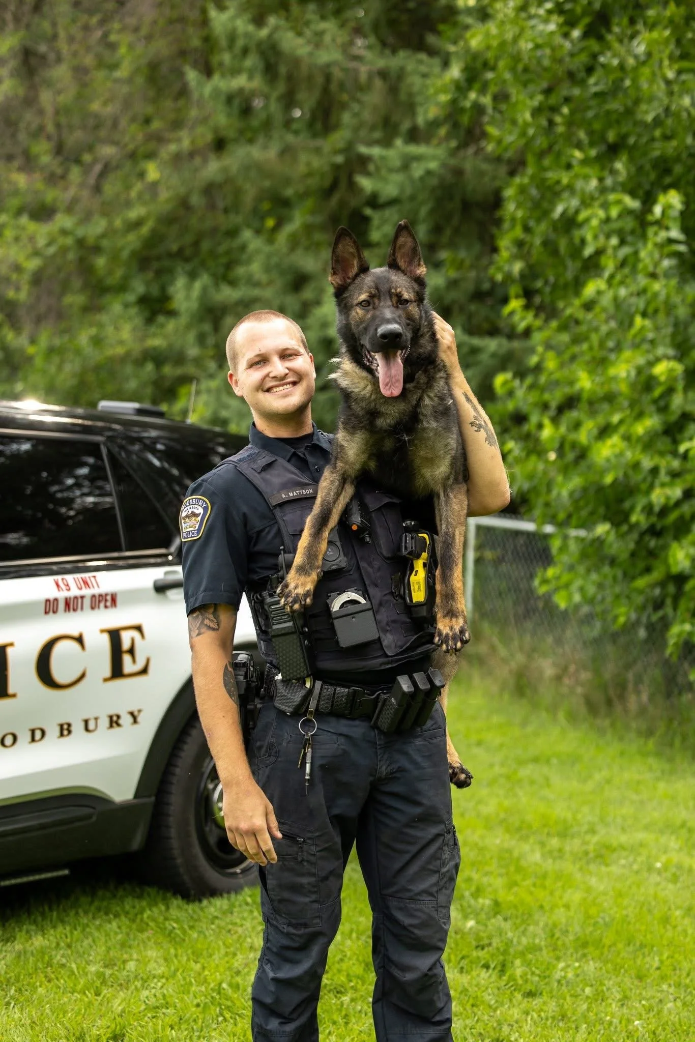 Police officer smiling while holding a German Shepherd dog on his shoulder outdoors with greenery in the background.