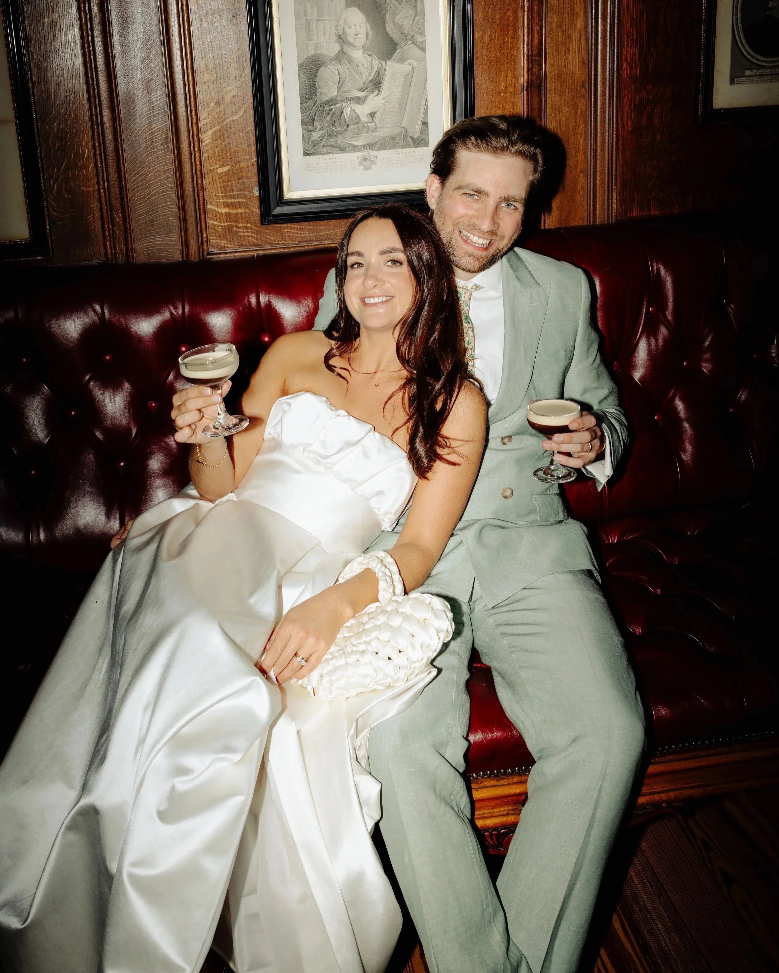 A smiling woman in a white dress and a smiling man in a light gray suit holding drinks, sitting on a red leather tufted bench in a wood-paneled room with framed pictures on the wall.