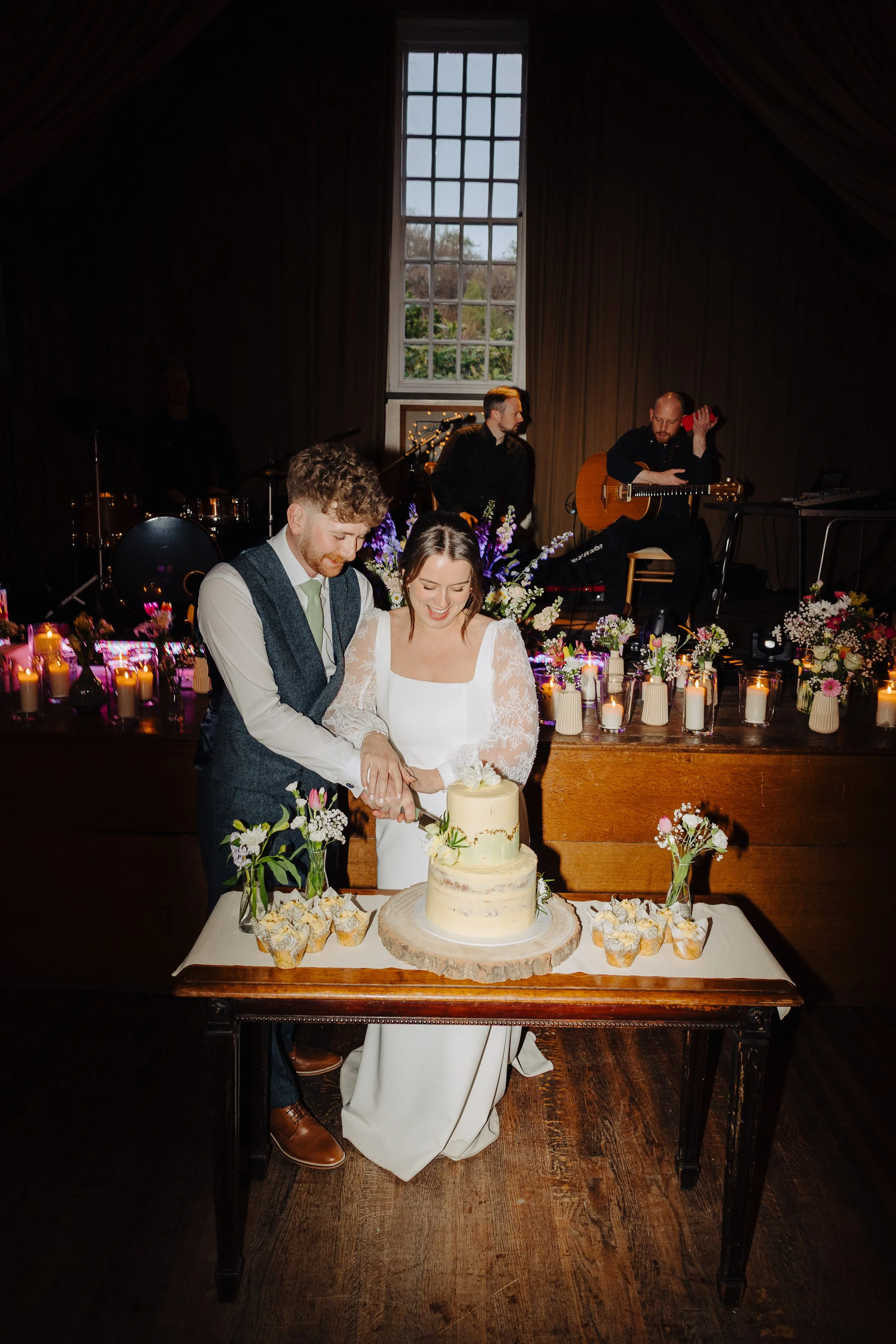 A bride and groom cutting a wedding cake at their reception. The cake is on a wooden table surrounded by flowers and candles. In the background, a band is performing with musicians playing instruments, including a guitar and drums, in a dimly lit roo