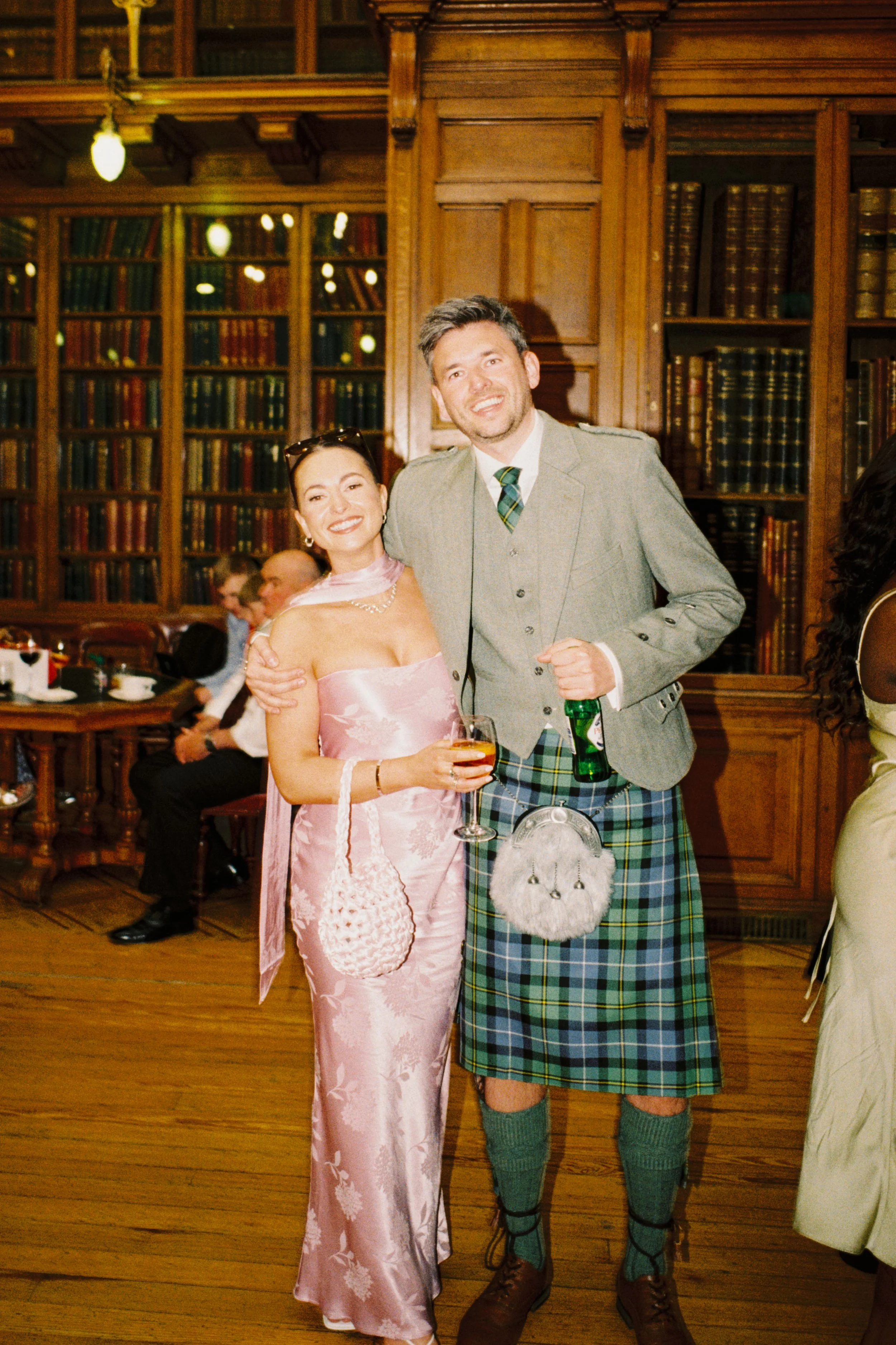 A woman in a pink satin dress holding a drink and a man in a traditional Scottish kilt outfit standing together in a wood-paneled room with bookshelves and other guests in the background.