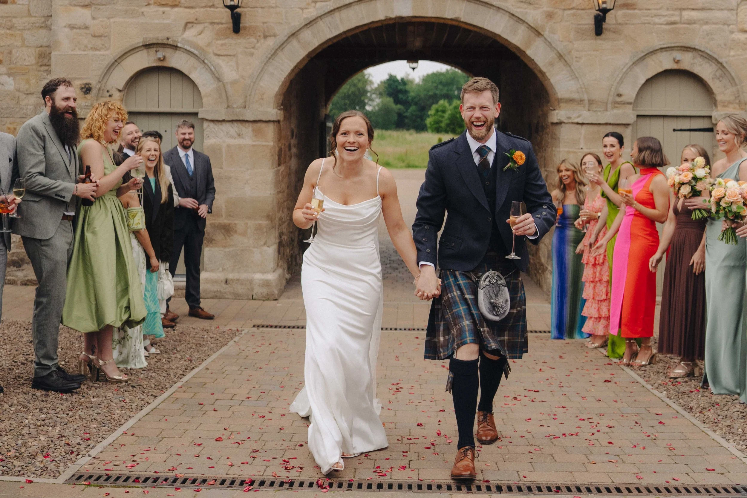 A bride and groom walking hand in hand down an aisle, smiling and laughing, surrounded by guests at a wedding ceremony outdoors with a stone archway in the background.