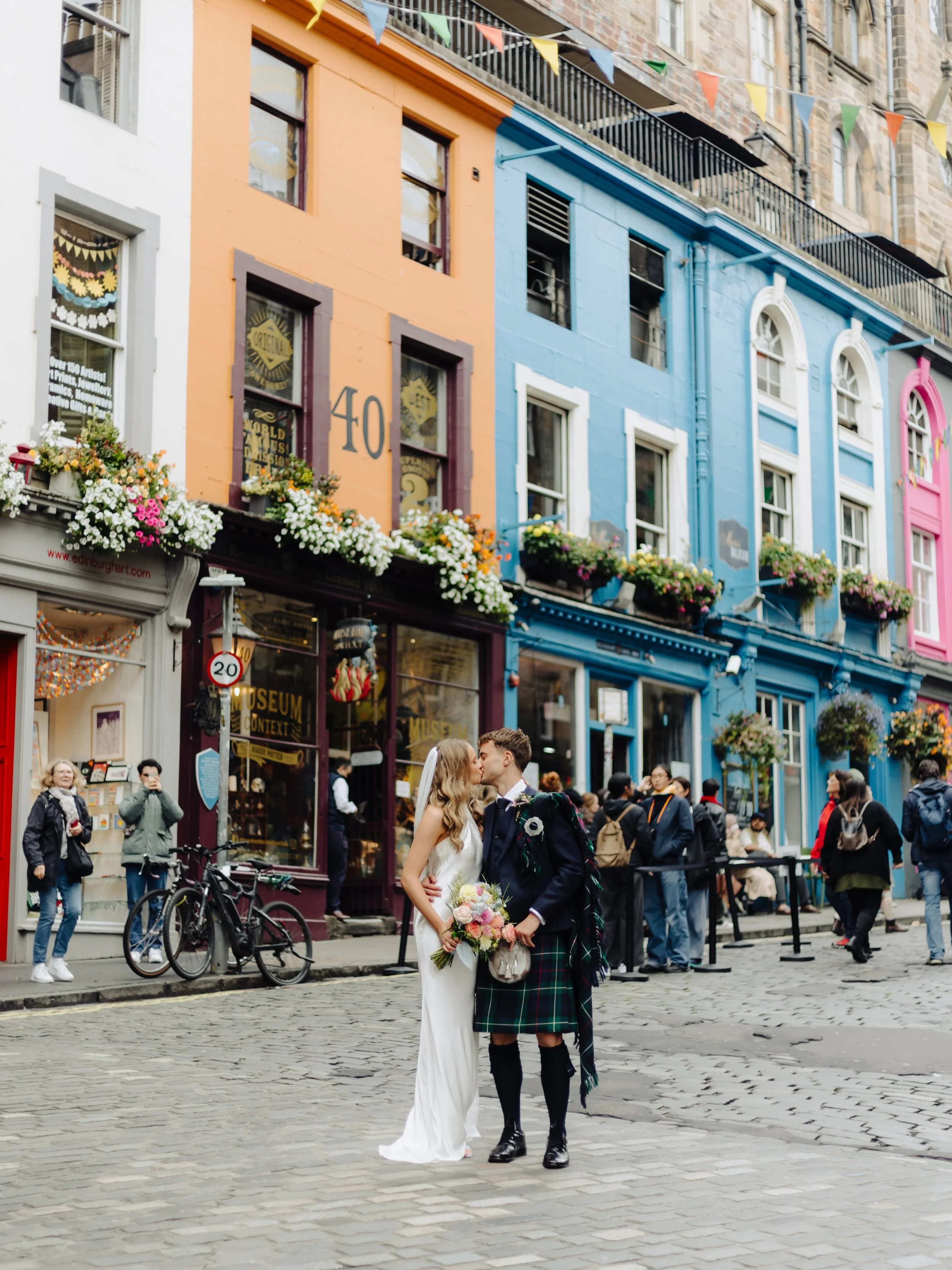 A newlywed couple sharing a kiss in front of colorful buildings on Cockburn street in Edinburgh City Scotland, with onlookers and shop fronts in the background.