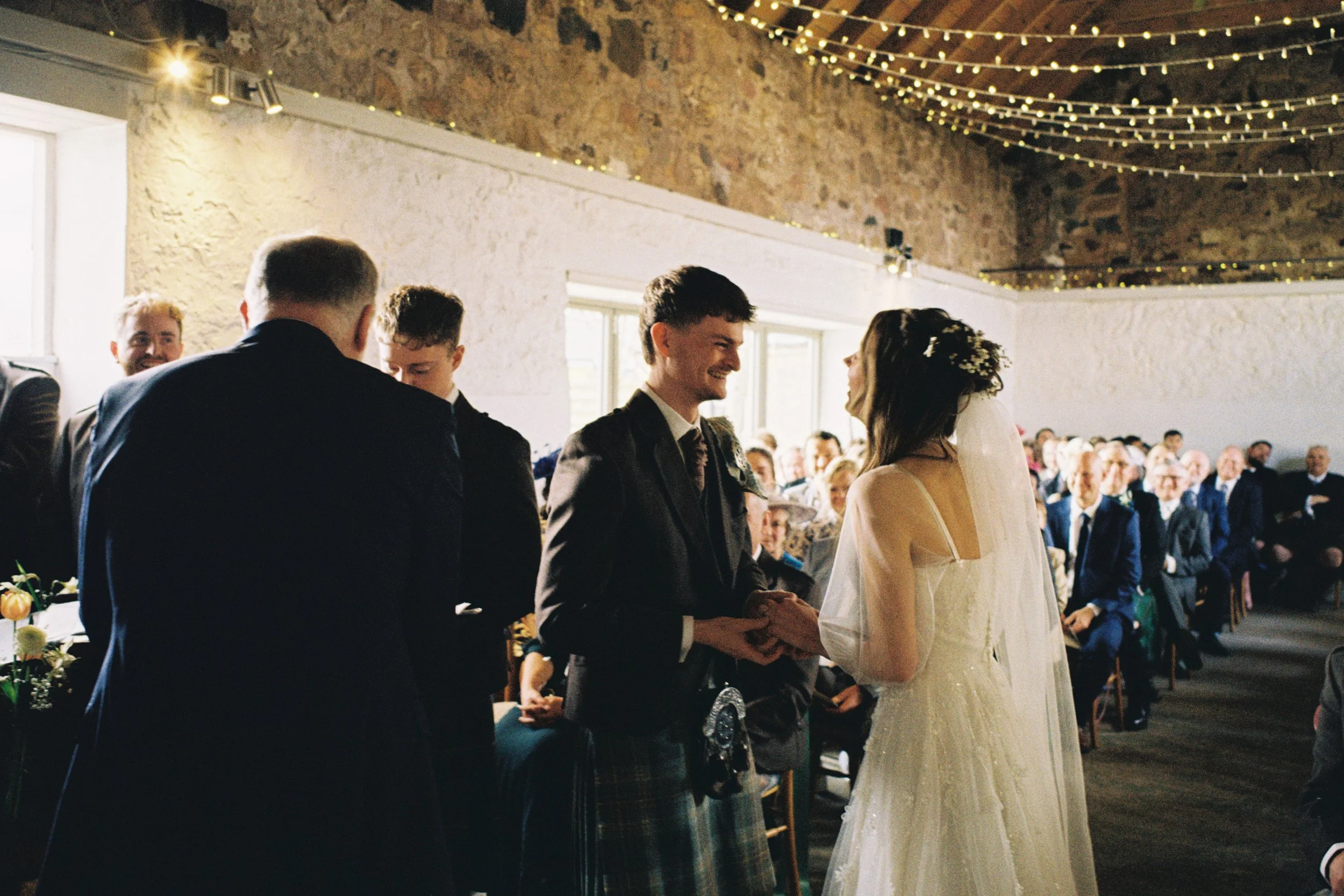 A wedding ceremony inside a rustic venue with string lights hanging from the ceiling. The bride and groom are facing each other, holding hands, with the officiant standing nearby. Guests are seated in the background, attentively watching the ceremony.