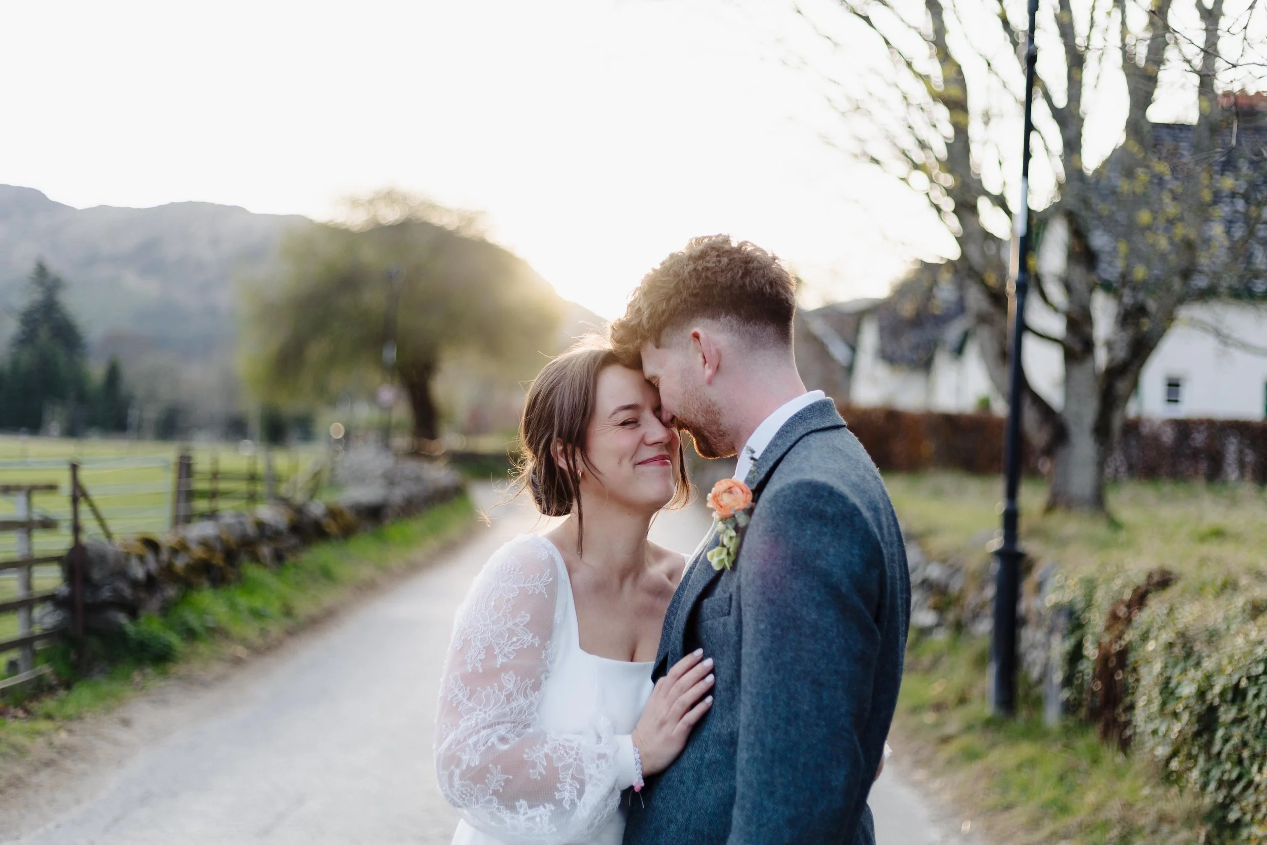A bride and groom share an intimate moment outside on a sunny day, with the bride smiling and touching the groom's chest, and the groom pressing his forehead against hers. The background includes trees, a wooden fence, and a pathway.