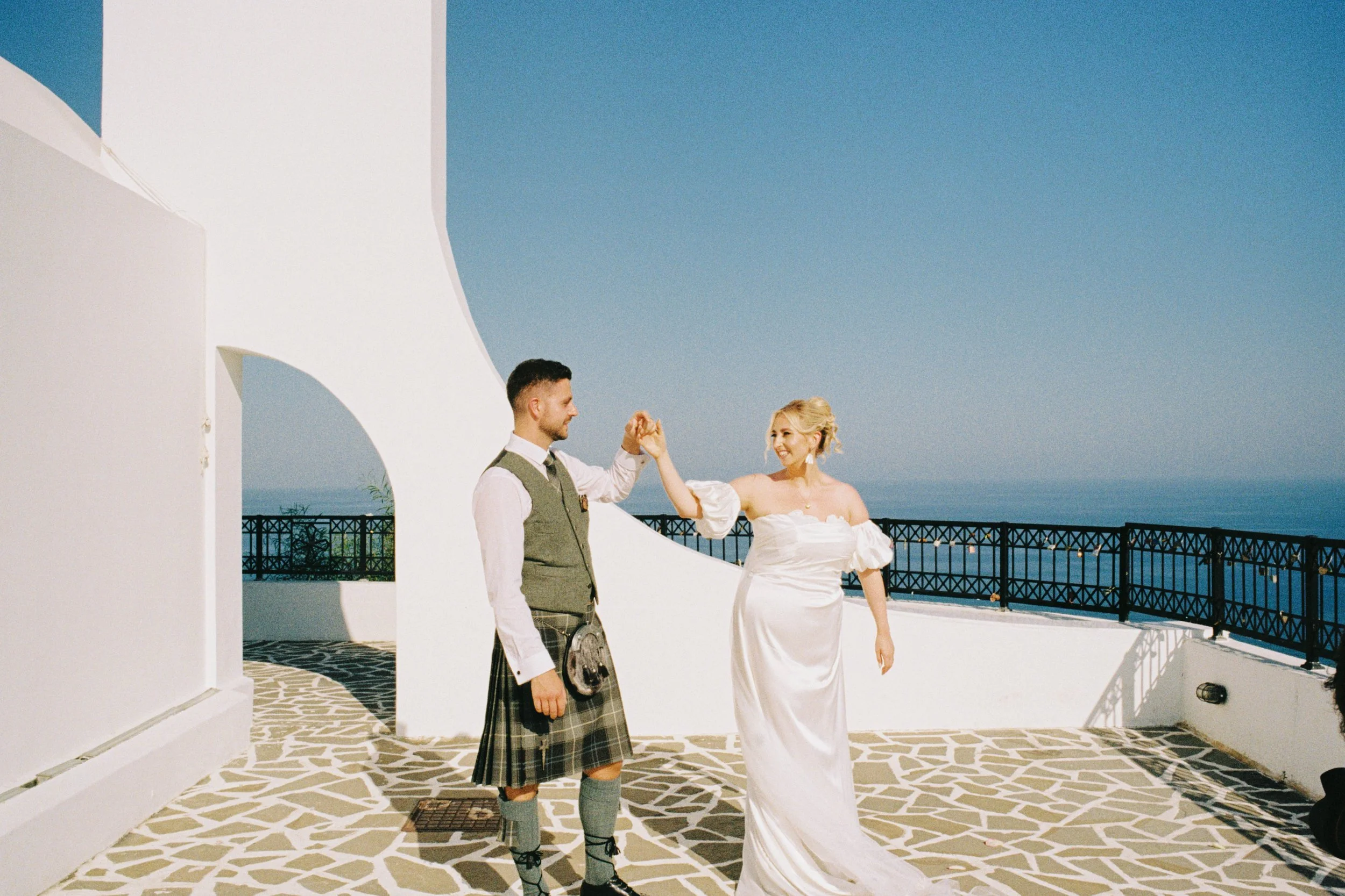A man and woman in wedding attire dance on a white balcony with a ocean view. The man wears a traditional Scottish kilt; the woman wears a white wedding dress. The sky is clear and blue.