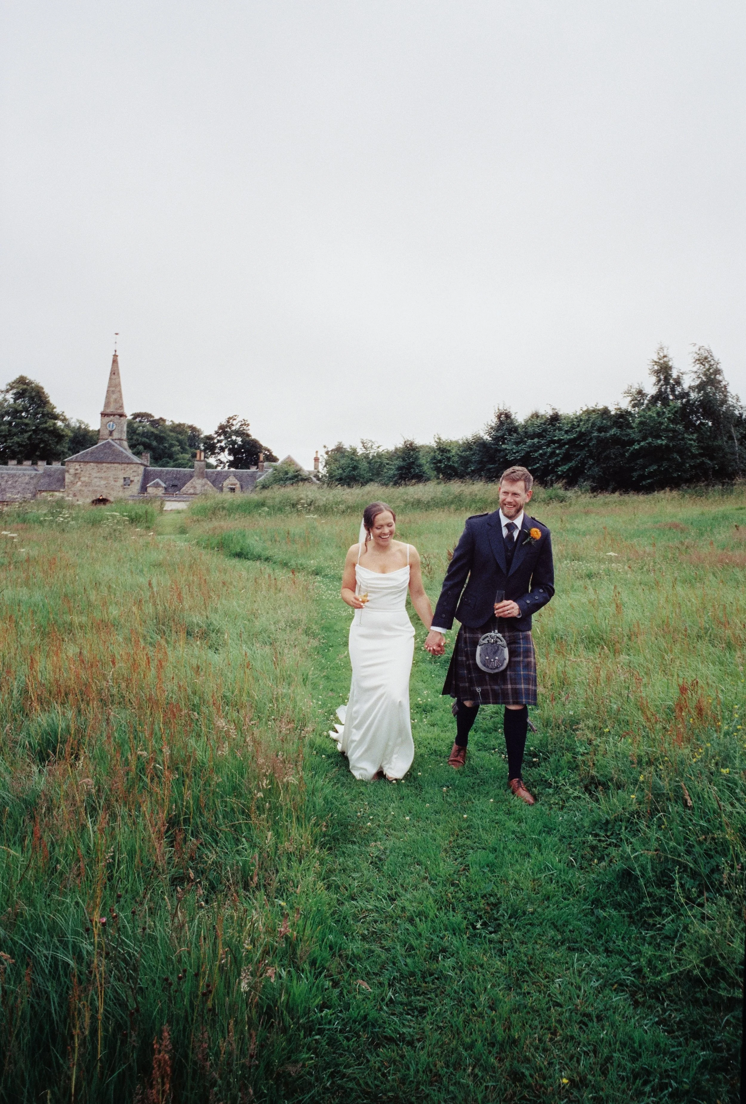 A bride and groom walking hand in hand through a grassy field on their wedding day, with a church and houses in the background.