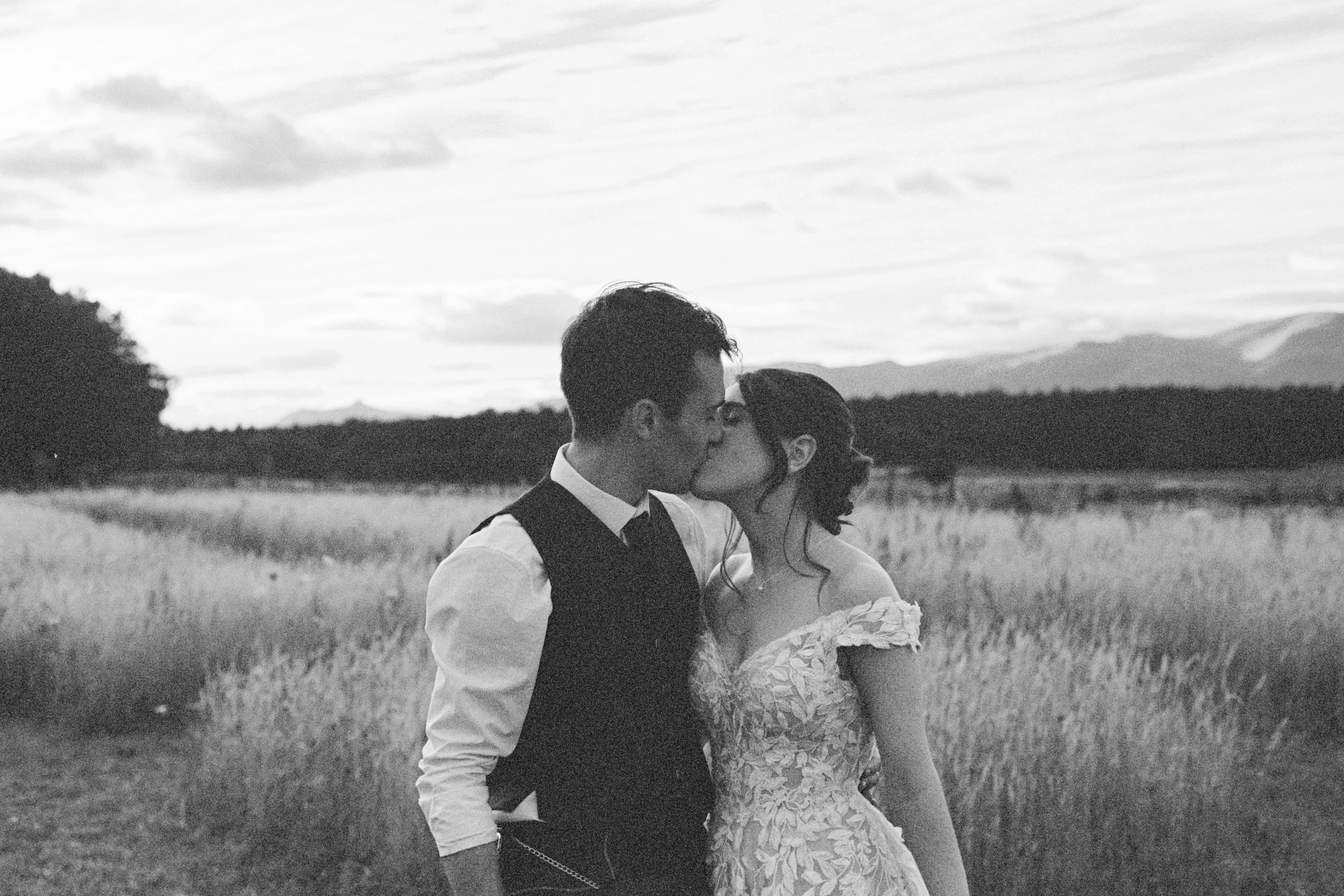 A black and white photo of a couple kissing in an open field, with mountains and clouds in the background.