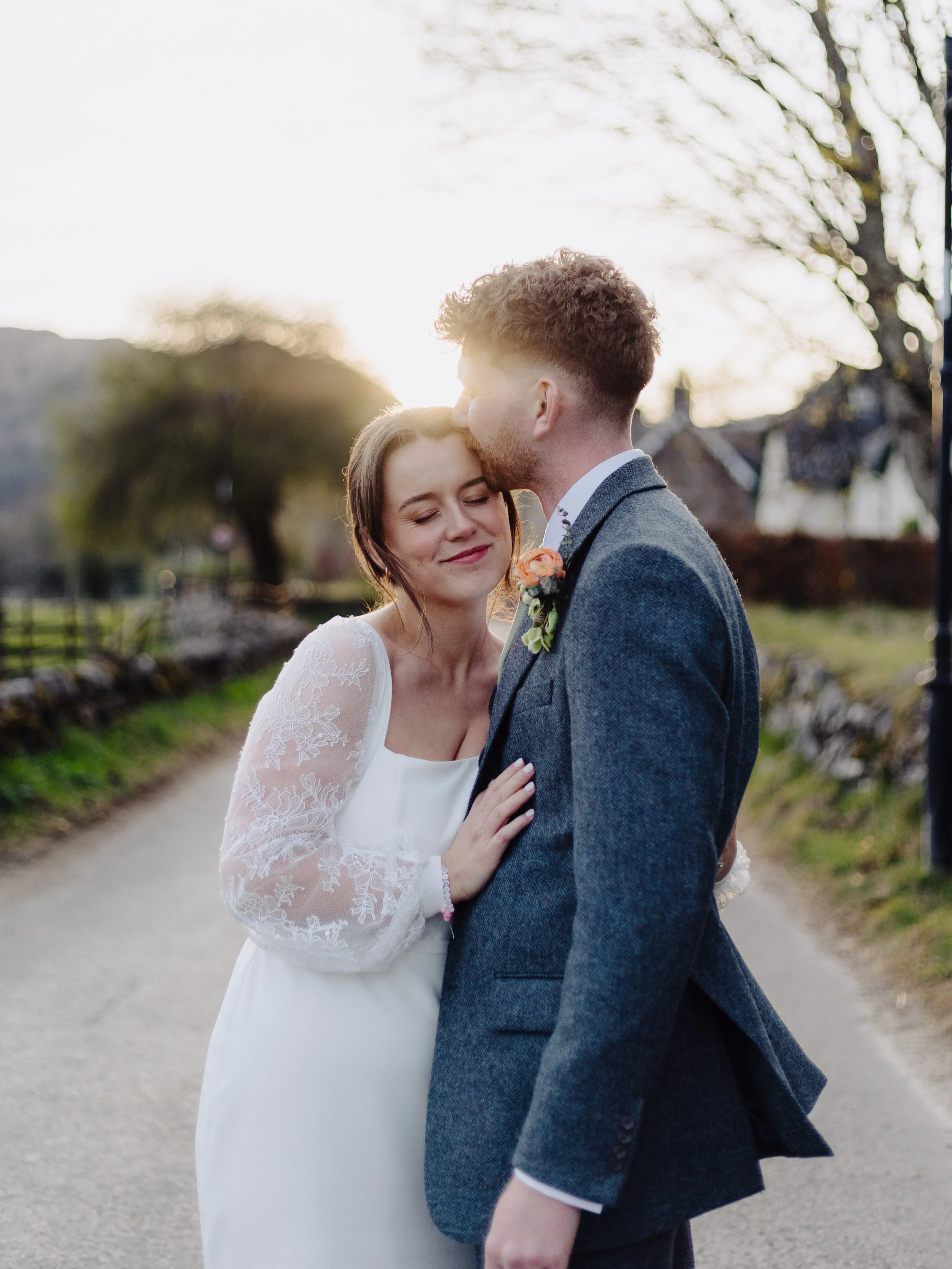 A bride and groom share a kiss outdoors at sunset, with the groom kissing the bride's forehead, and the bride smiling with eyes closed, wearing a white lace wedding dress, and the groom in a gray suit with a boutonniere in a natural setting.