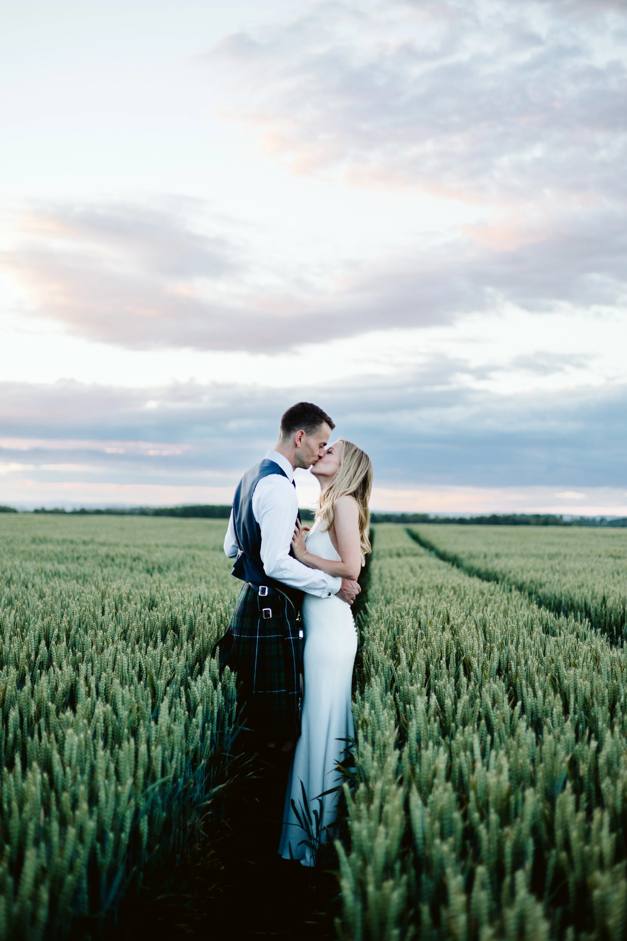A newly married couple in wedding attire sharing a kiss in a green wheat field with a cloudy sky overhead.