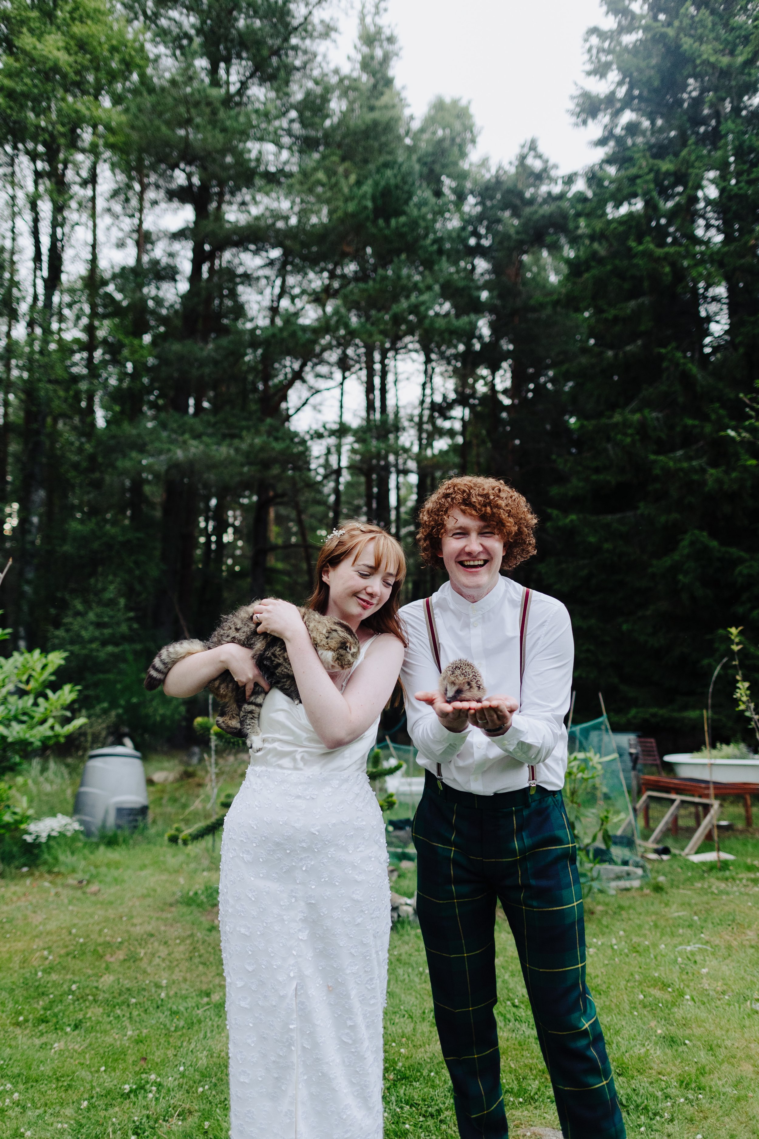 A woman in a white dress holding a kitten, and a man in a white shirt and plaid pants holding a kitten, standing outdoors in a grassy backyard with trees.